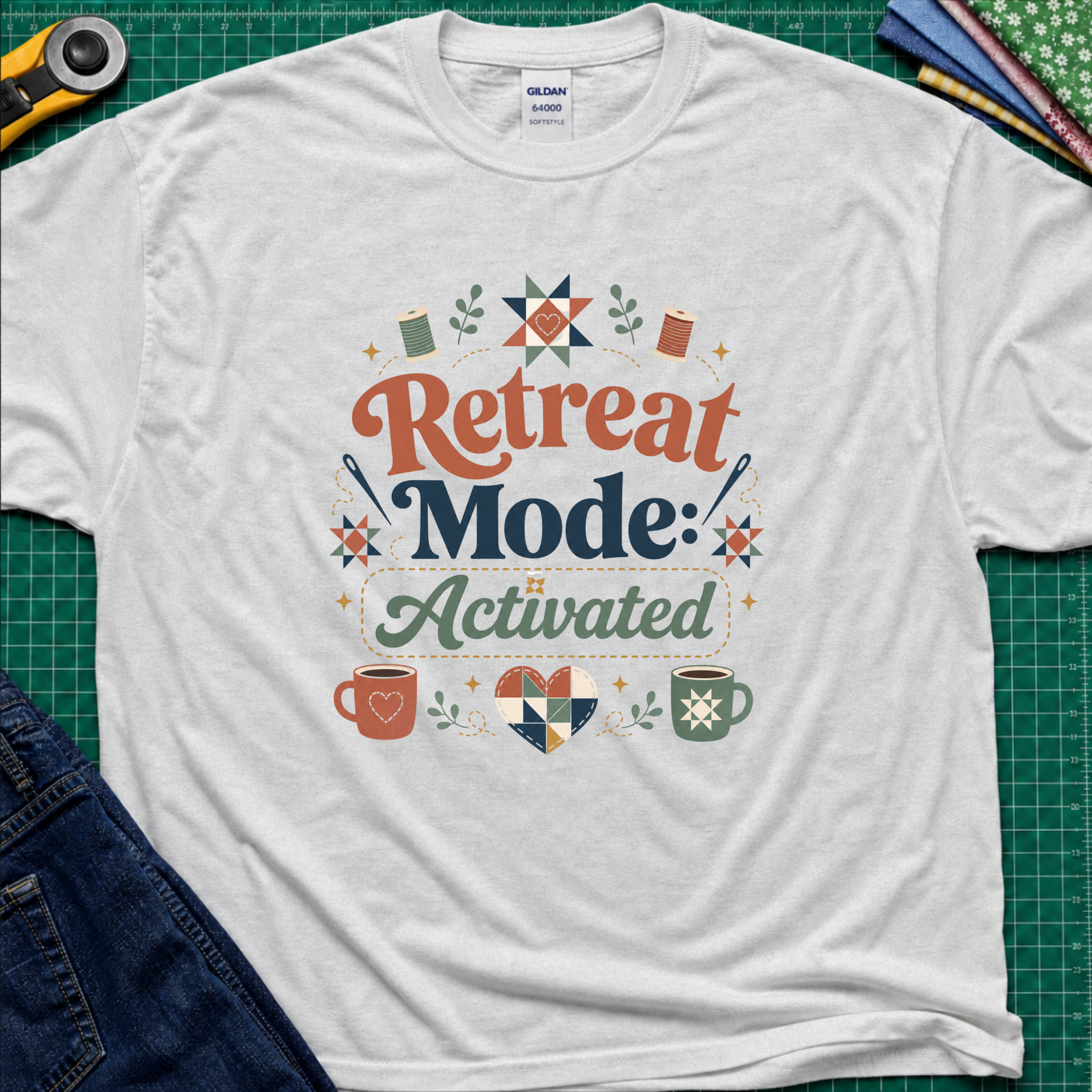 A light grey Gildan t-shirt with a colorful retro graphic that reads 'Retreat Mode: Activated' surrounded by quilting and coffee-themed illustrations. The shirt is displayed flat on a green cutting mat with a rotary cutter, blue jeans and fabric partially visible in the corners.