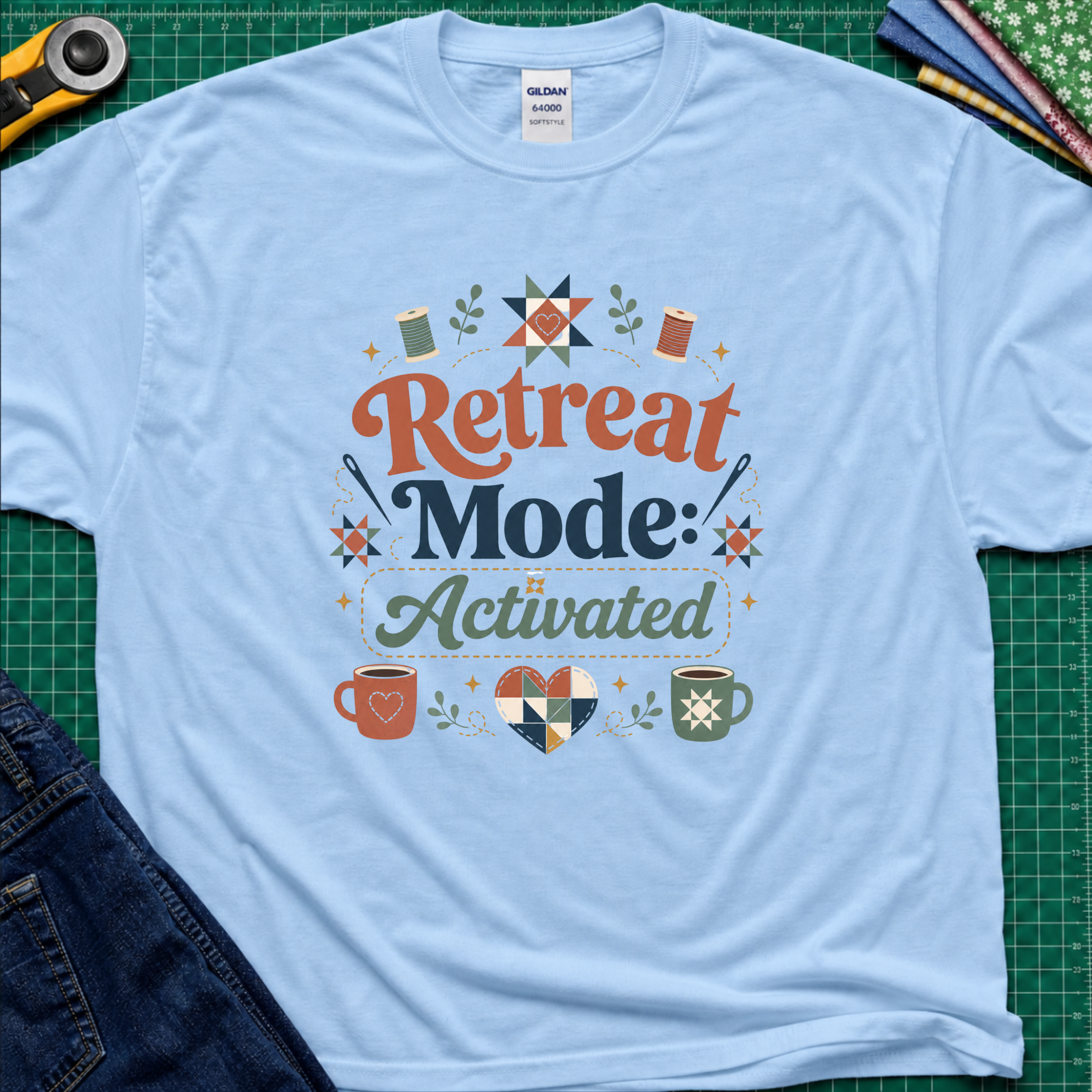 A light blue Gildan t-shirt with a colorful retro graphic that reads 'Retreat Mode: Activated' surrounded by quilting and coffee-themed illustrations. The shirt is displayed flat on a green cutting mat with a rotary cutter, blue jeans and fabric partially visible in the corners.