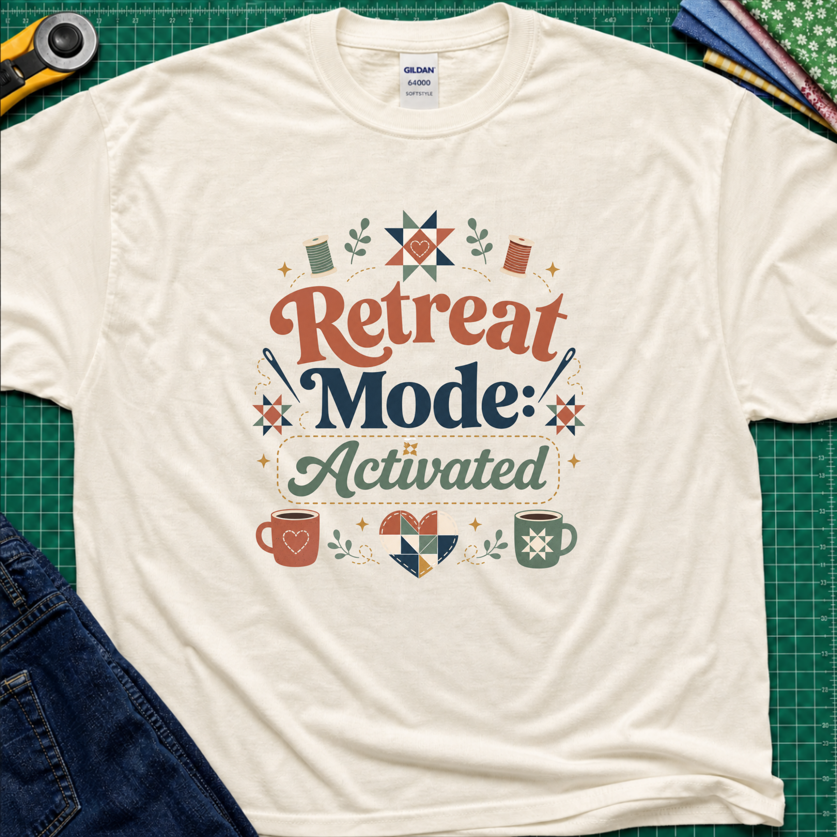 A light cream Gildan t-shirt with a colorful retro graphic that reads 'Retreat Mode: Activated' surrounded by quilting and coffee-themed illustrations. The shirt is displayed flat on a green cutting mat with a rotary cutter, blue jeans and fabric partially visible in the corners.