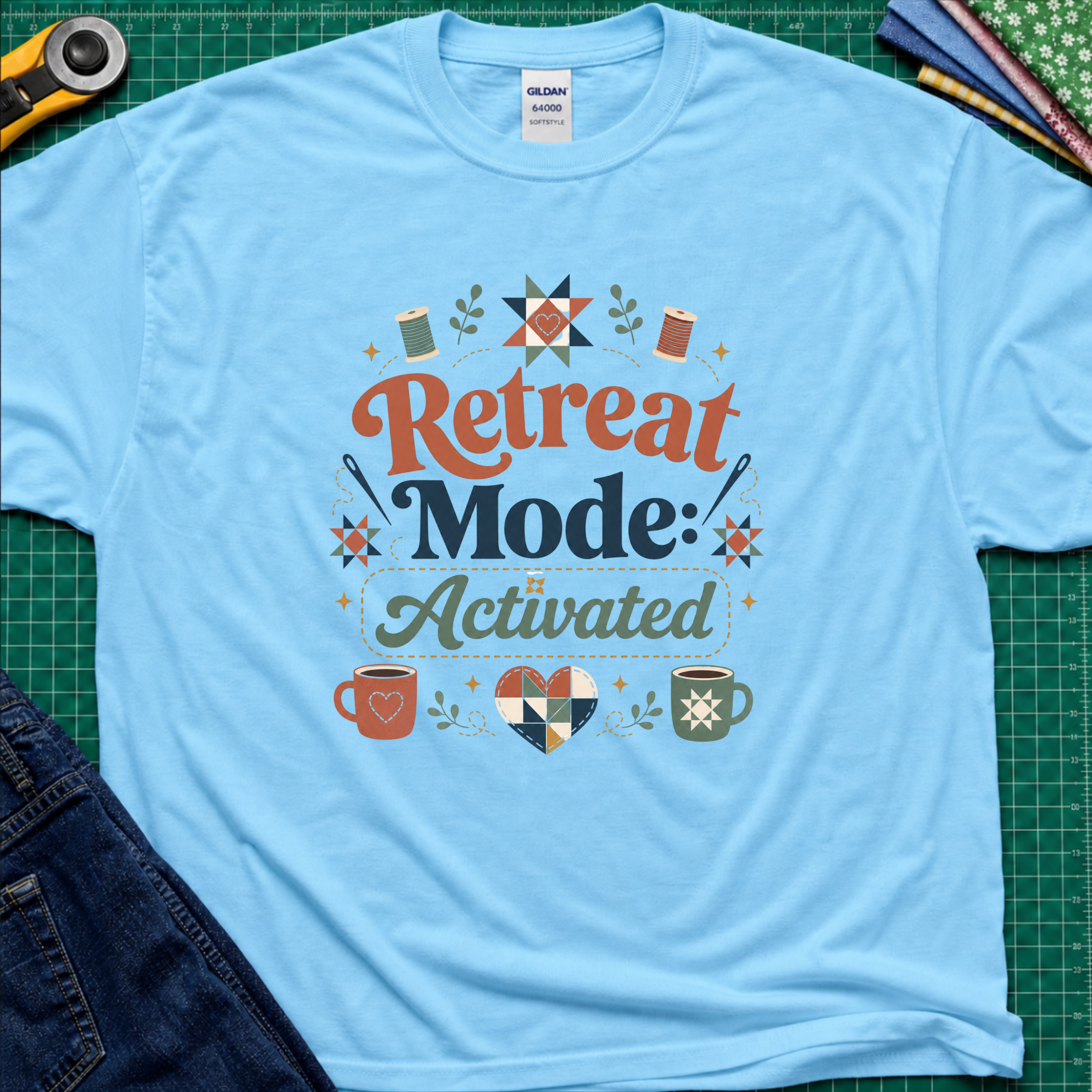 A sky blue Gildan t-shirt with a colorful retro graphic that reads 'Retreat Mode: Activated' surrounded by quilting and coffee-themed illustrations. The shirt is displayed flat on a green cutting mat with a rotary cutter, blue jeans and fabric partially visible in the corners.
