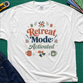 A white Gildan t-shirt with a colorful retro graphic that reads 'Retreat Mode: Activated' surrounded by quilting and coffee-themed illustrations. The shirt is displayed flat on a green cutting mat with a rotary cutter, blue jeans and fabric partially visible in the corners.