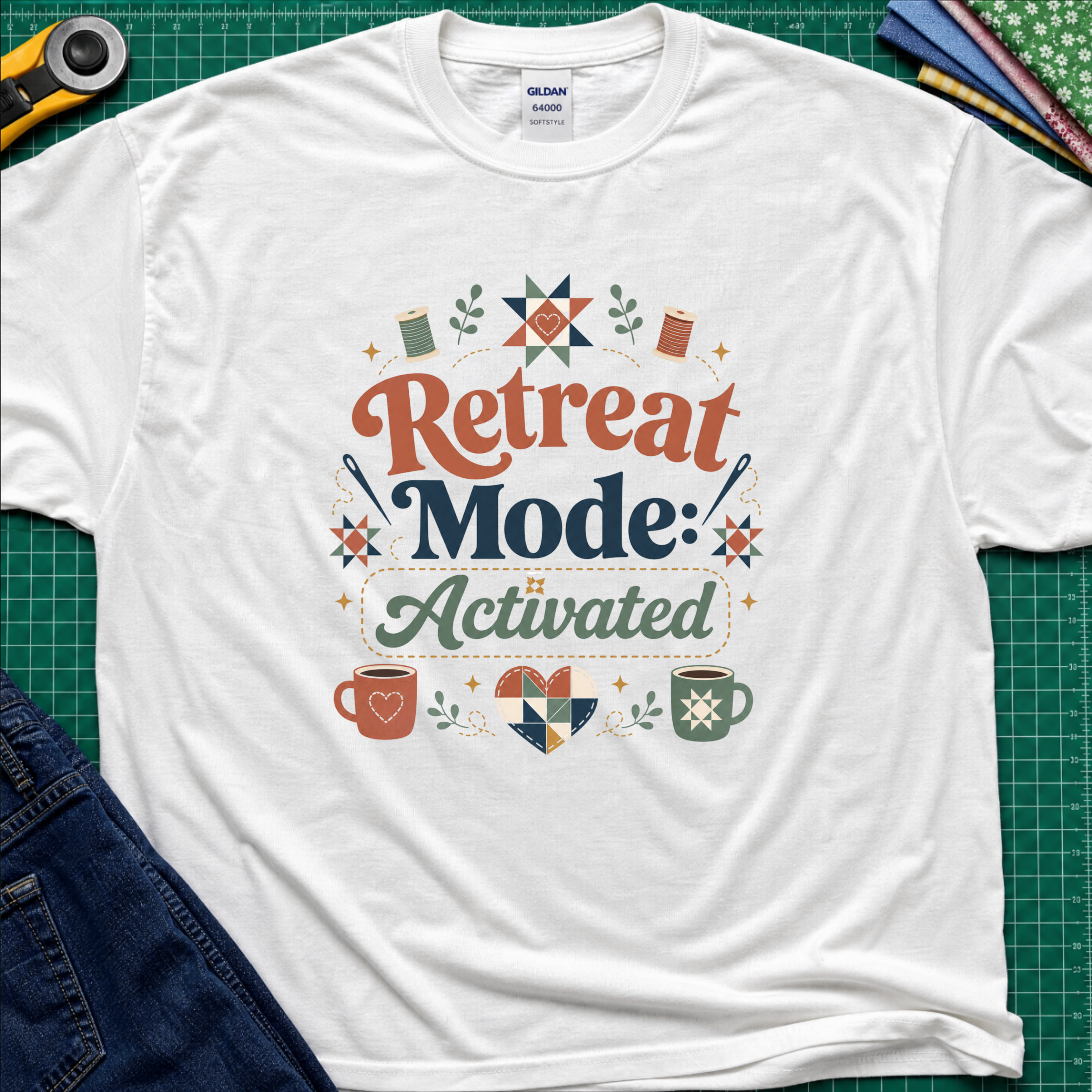A white Gildan t-shirt with a colorful retro graphic that reads 'Retreat Mode: Activated' surrounded by quilting and coffee-themed illustrations. The shirt is displayed flat on a green cutting mat with a rotary cutter, blue jeans and fabric partially visible in the corners.