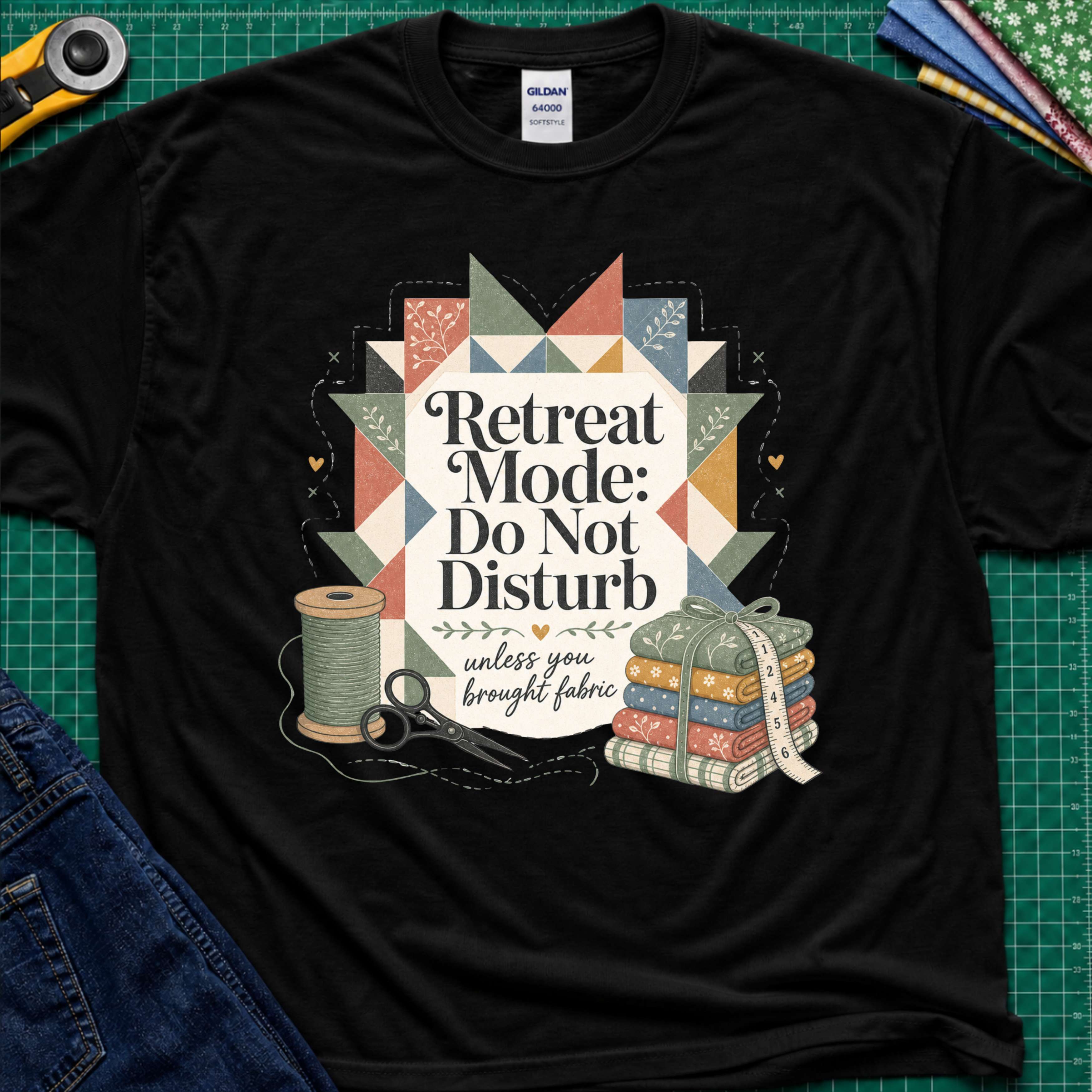 A black Gildan t-shirt with a retro quilted patchwork graphic and the text 'Retreat Mode: Do Not Disturb unless you brought fabric', laid flat on a green cutting mat with sewing props.