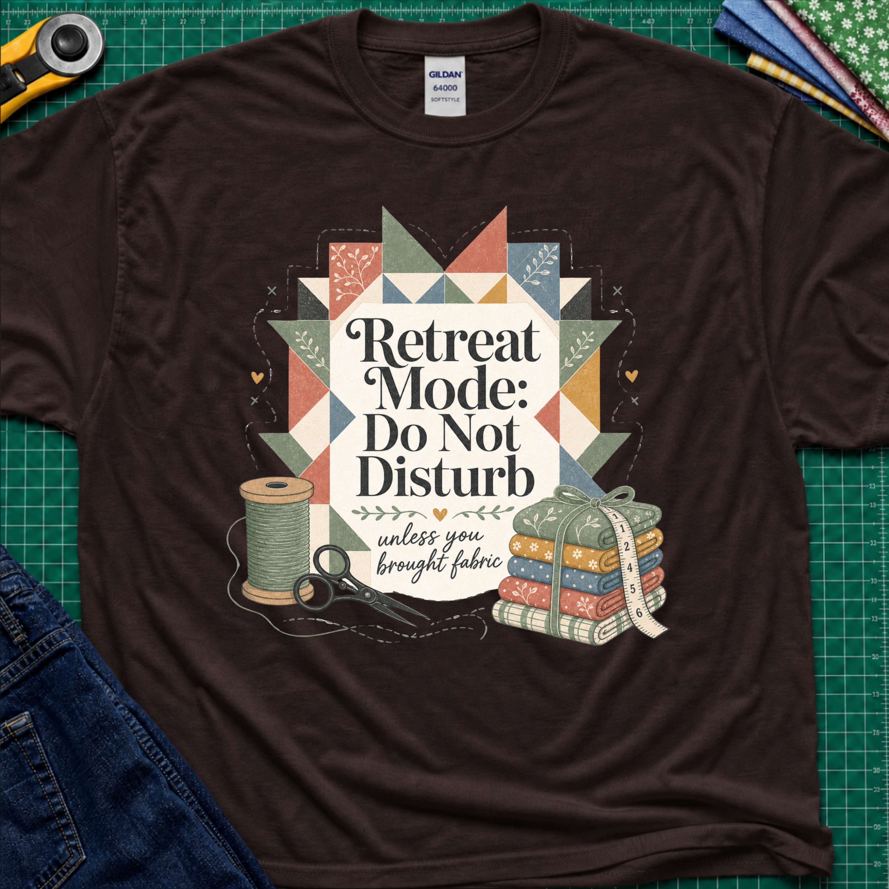 A chocolate brown Gildan t-shirt with a retro quilted patchwork graphic and the text 'Retreat Mode: Do Not Disturb unless you brought fabric', laid flat on a green cutting mat with sewing props.