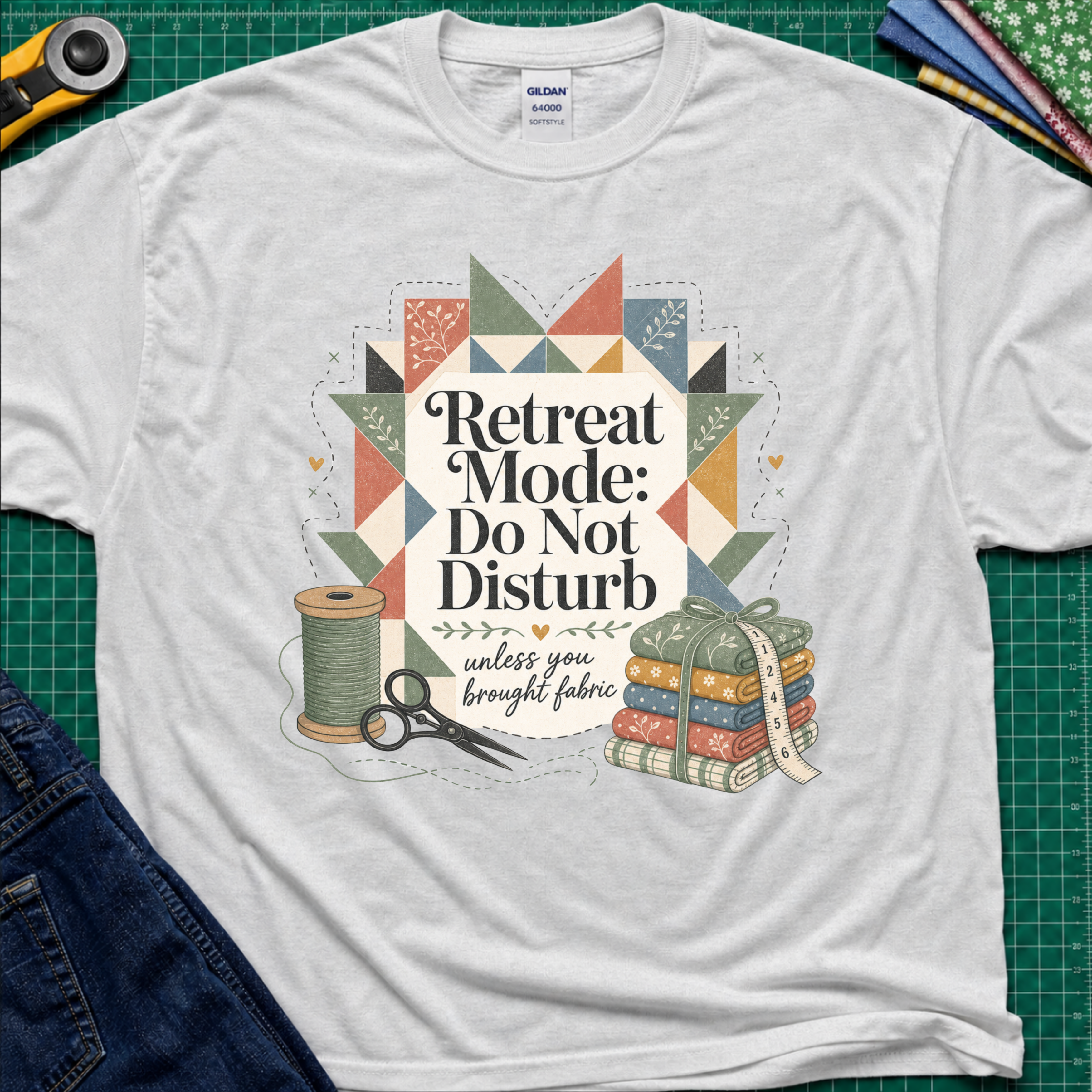 A light grey Gildan t-shirt with a retro quilted patchwork graphic and the text 'Retreat Mode: Do Not Disturb unless you brought fabric', laid flat on a green cutting mat with sewing props.