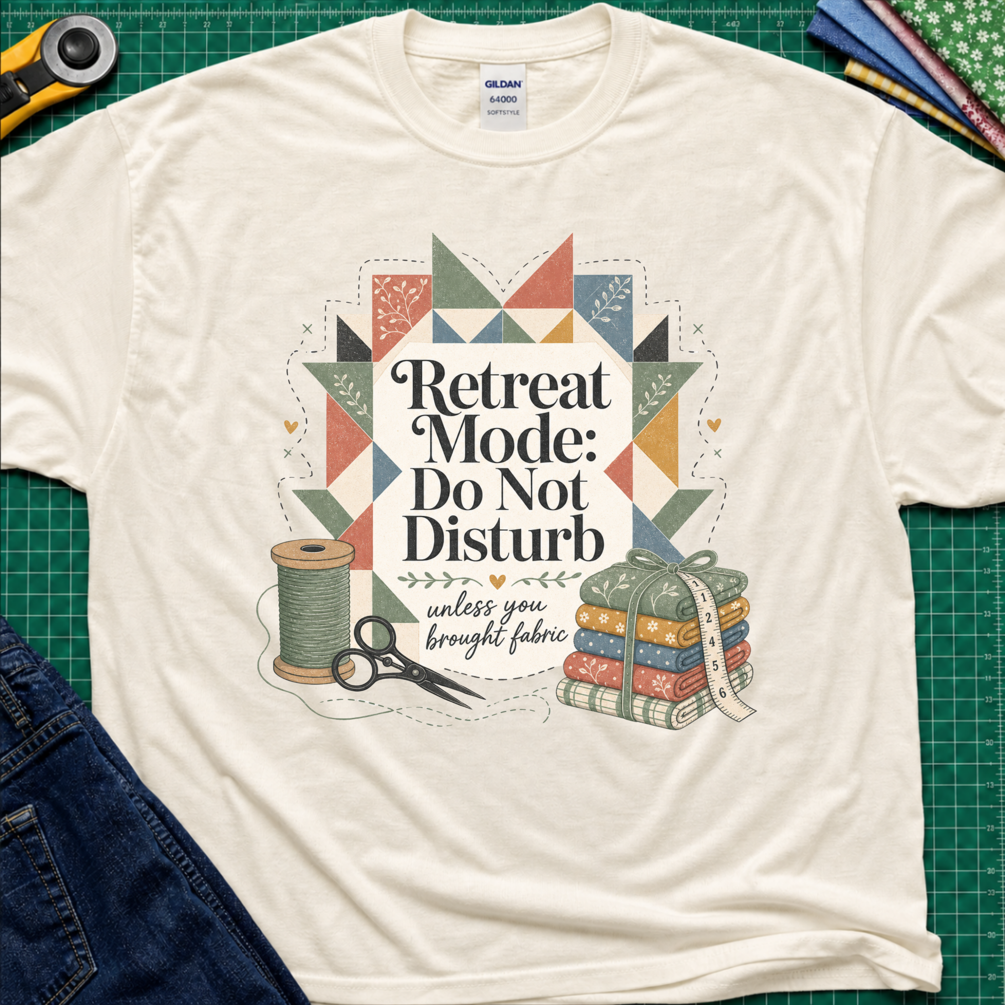 A light cream Gildan t-shirt with a retro quilted patchwork graphic and the text 'Retreat Mode: Do Not Disturb unless you brought fabric', laid flat on a green cutting mat with sewing props.