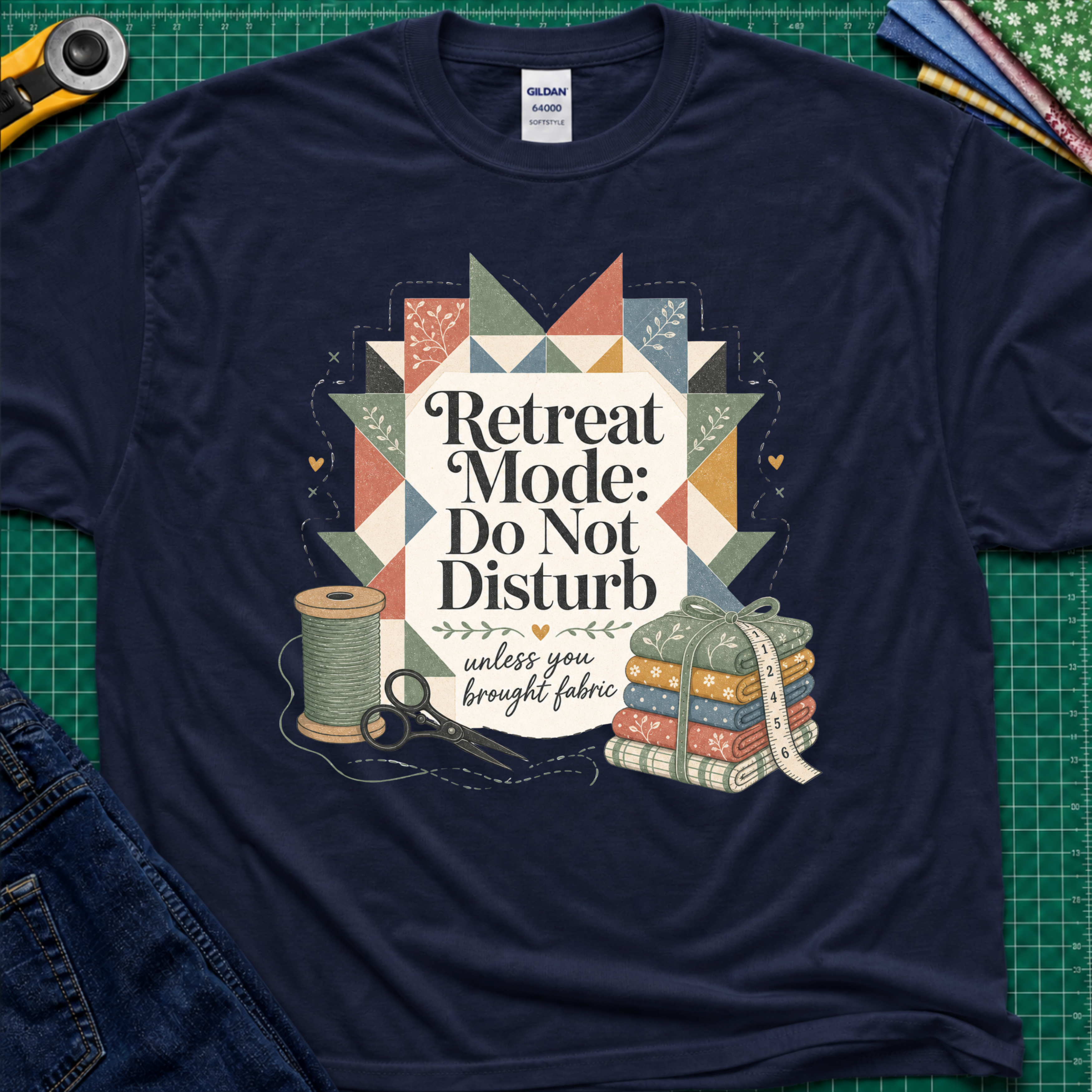 A navy blue Gildan t-shirt with a retro quilted patchwork graphic and the text 'Retreat Mode: Do Not Disturb unless you brought fabric', laid flat on a green cutting mat with sewing props.