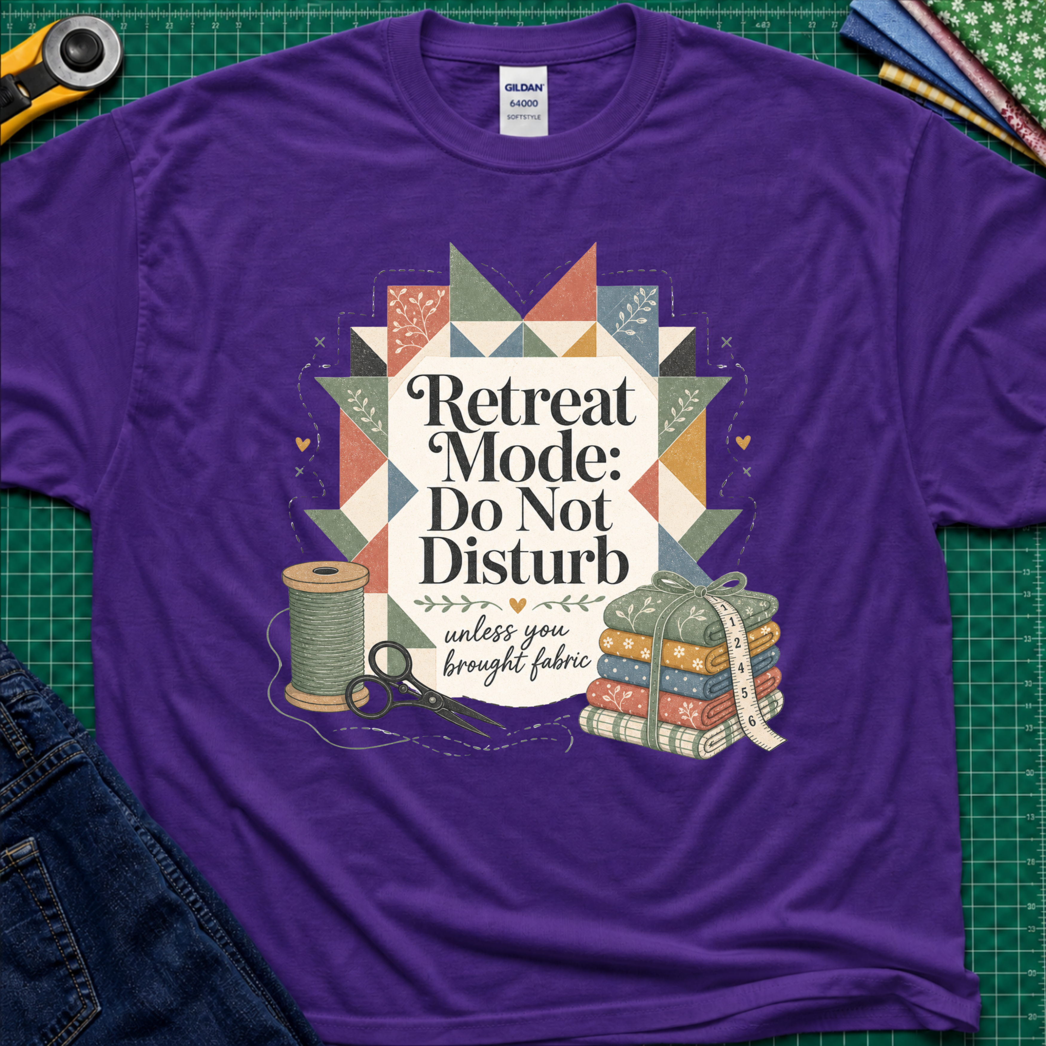 A purple Gildan t-shirt with a retro quilted patchwork graphic and the text 'Retreat Mode: Do Not Disturb unless you brought fabric', laid flat on a green cutting mat with sewing props.