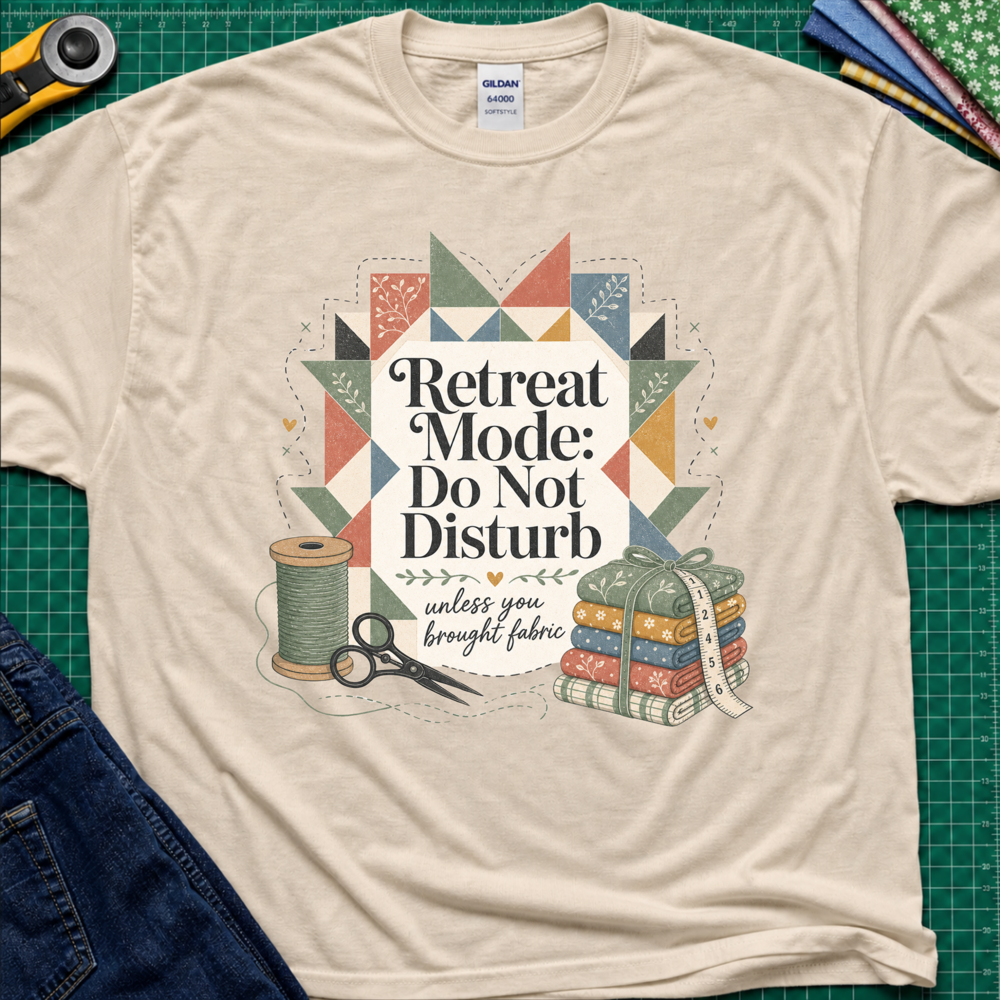 A dark cream Gildan t-shirt with a retro quilted patchwork graphic and the text 'Retreat Mode: Do Not Disturb unless you brought fabric', laid flat on a green cutting mat with sewing props.