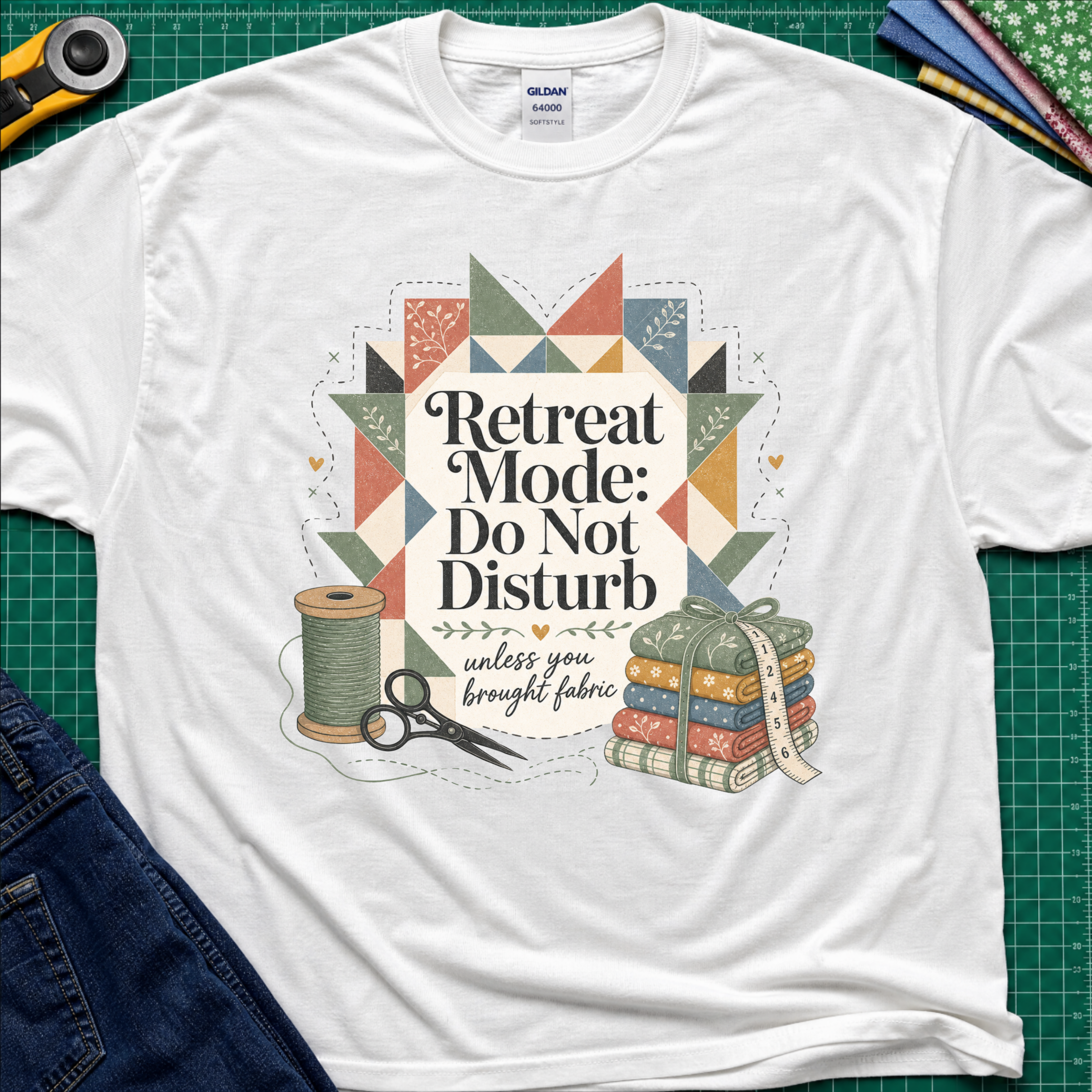 A white Gildan t-shirt with a retro quilted patchwork graphic and the text 'Retreat Mode: Do Not Disturb unless you brought fabric', laid flat on a green cutting mat with sewing props.