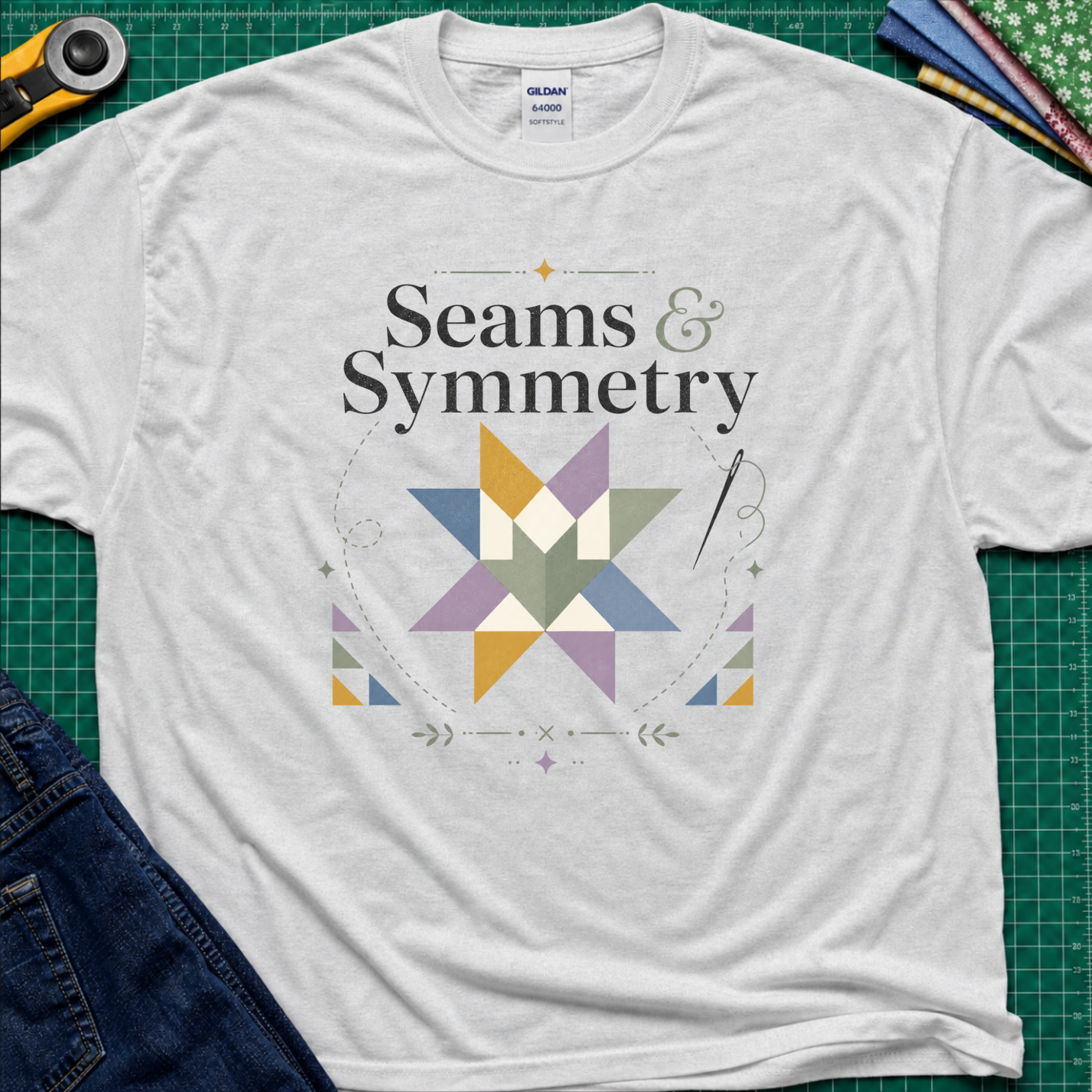 A light grey Gildan t-shirt with a 'Seams & Symmetry' graphic print with quilt block, needle and thread, laid flat on a green cutting mat with sewing props like a rotary cutter and denim jeans.
