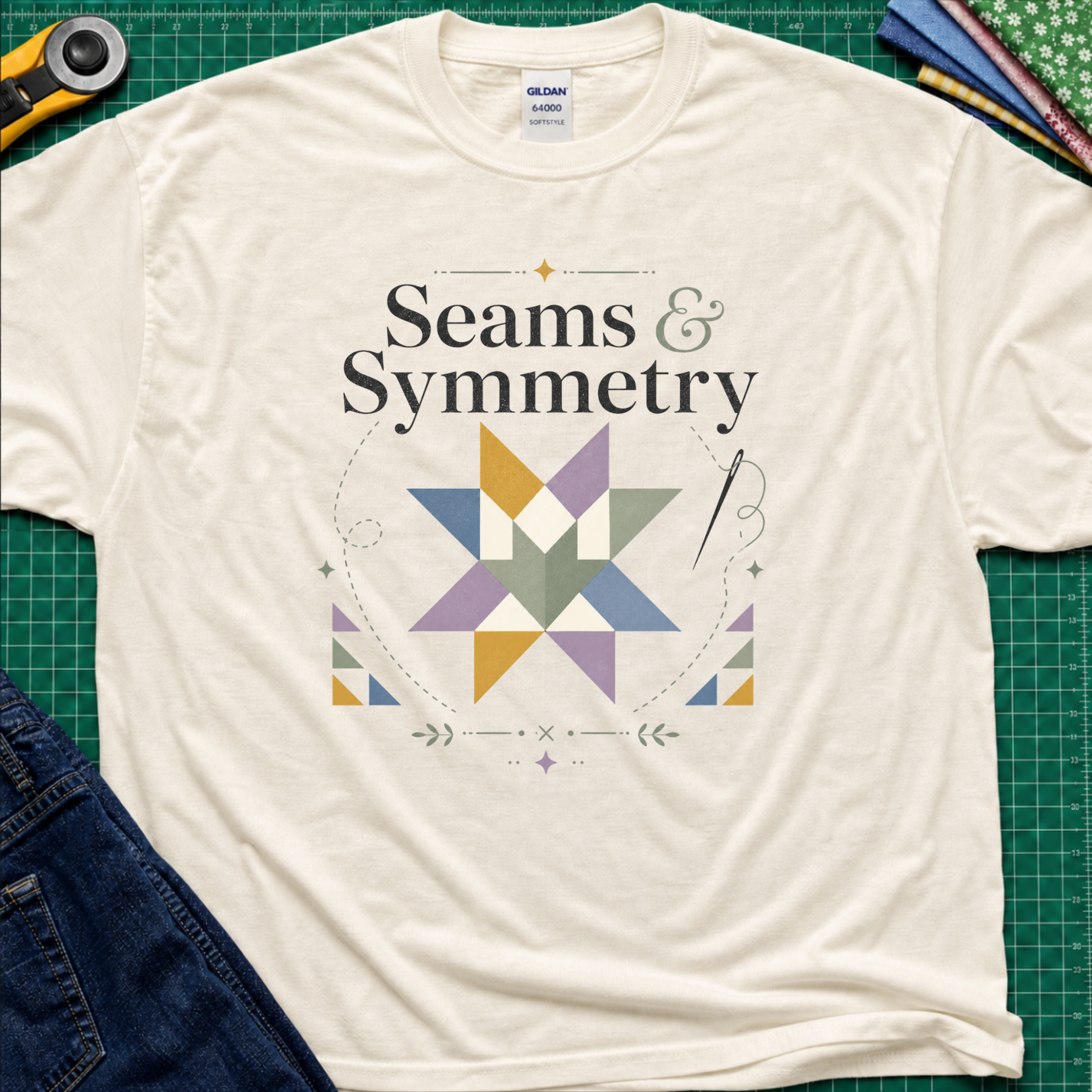 A light cream Gildan t-shirt with a 'Seams & Symmetry' graphic print with quilt block, needle and thread, laid flat on a green cutting mat with sewing props like a rotary cutter and denim jeans.