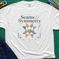 A white Gildan t-shirt with a 'Seams & Symmetry' graphic print with quilt block, needle and thread, laid flat on a green cutting mat with sewing props like a rotary cutter and denim jeans.