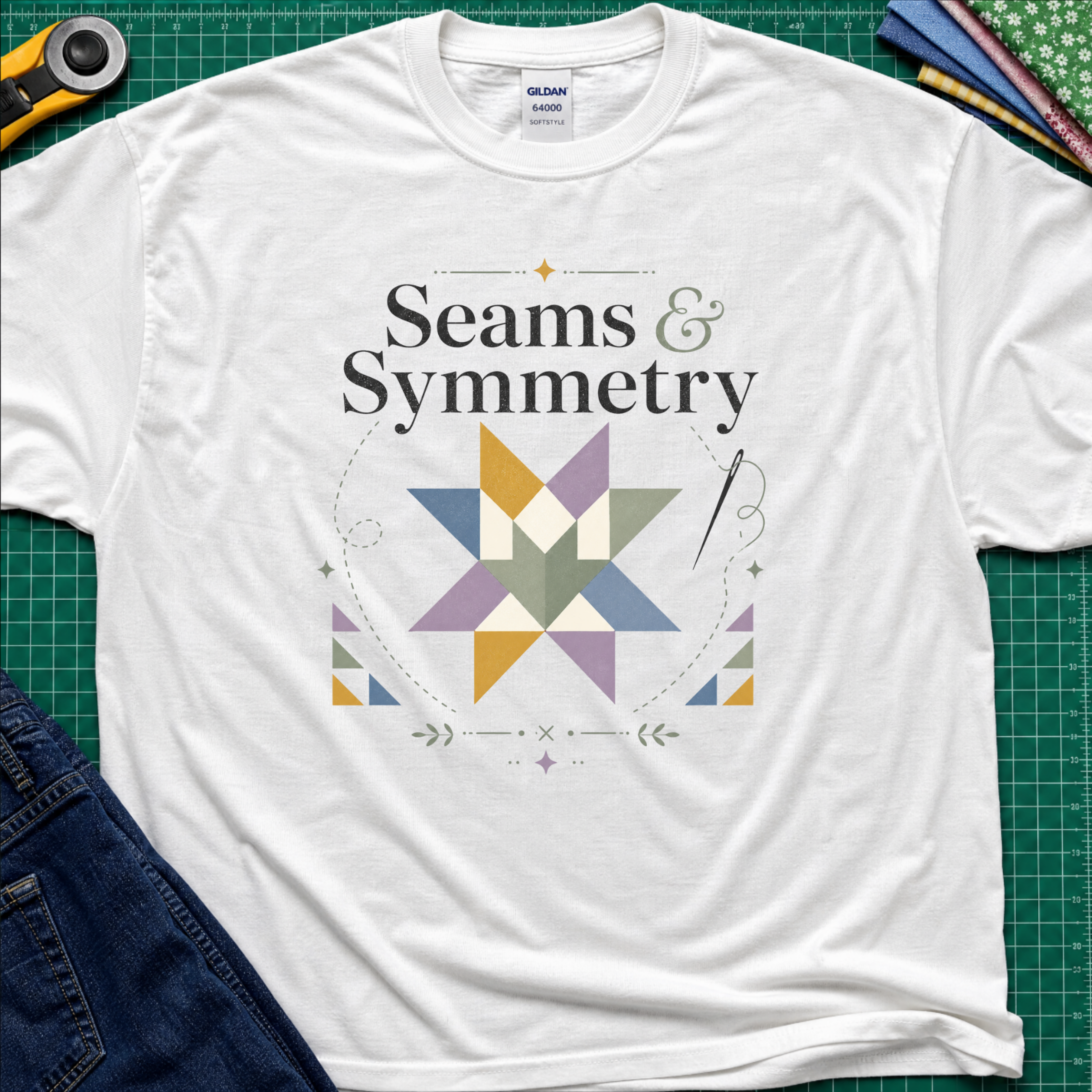 A white Gildan t-shirt with a 'Seams & Symmetry' graphic print with quilt block, needle and thread, laid flat on a green cutting mat with sewing props like a rotary cutter and denim jeans.