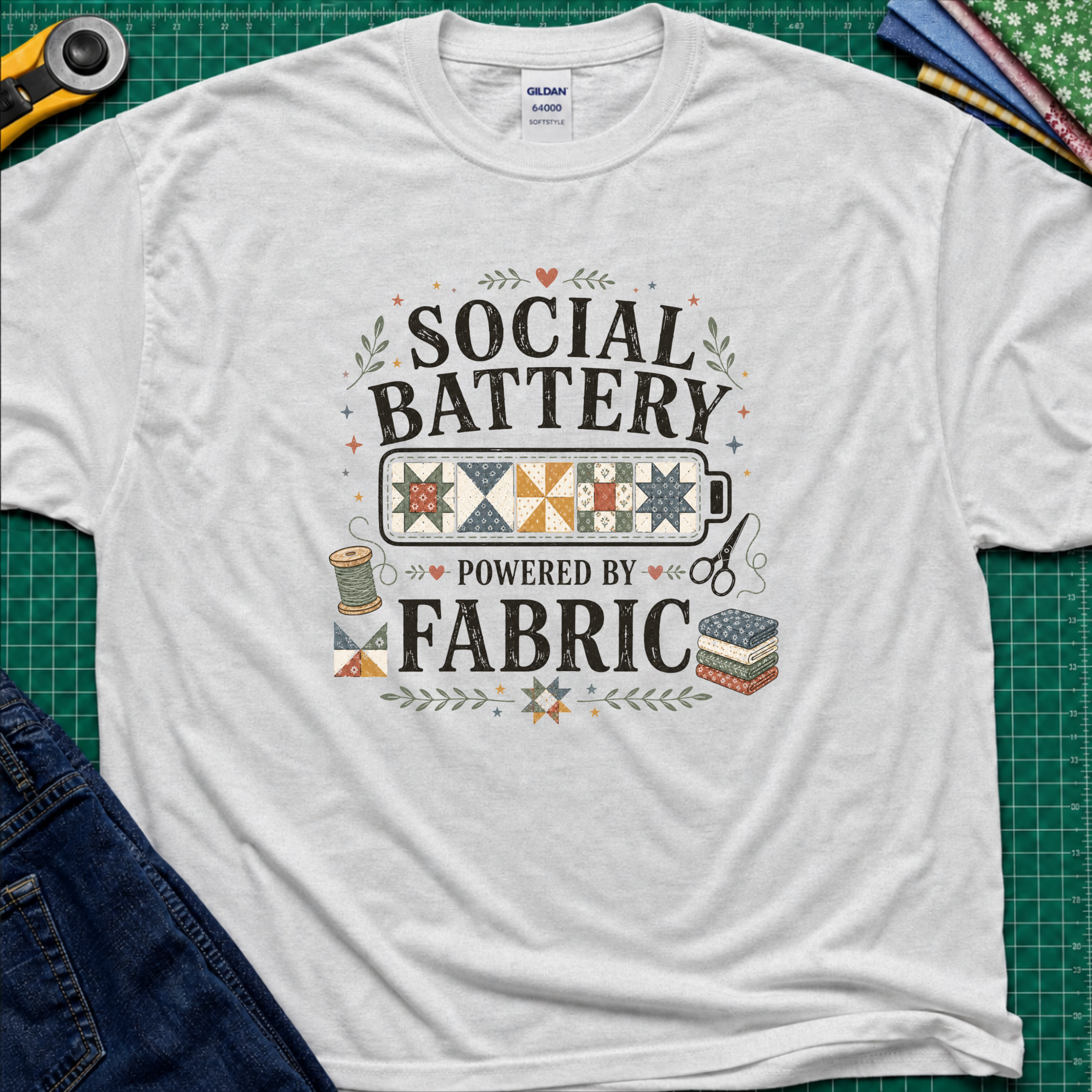 A light grey Gildan t-shirt with a 'Social Battery Powered by Fabric' graphic, laid flat on a green cutting mat with sewing props like a rotary cutter, scissors, and fabric rolls.