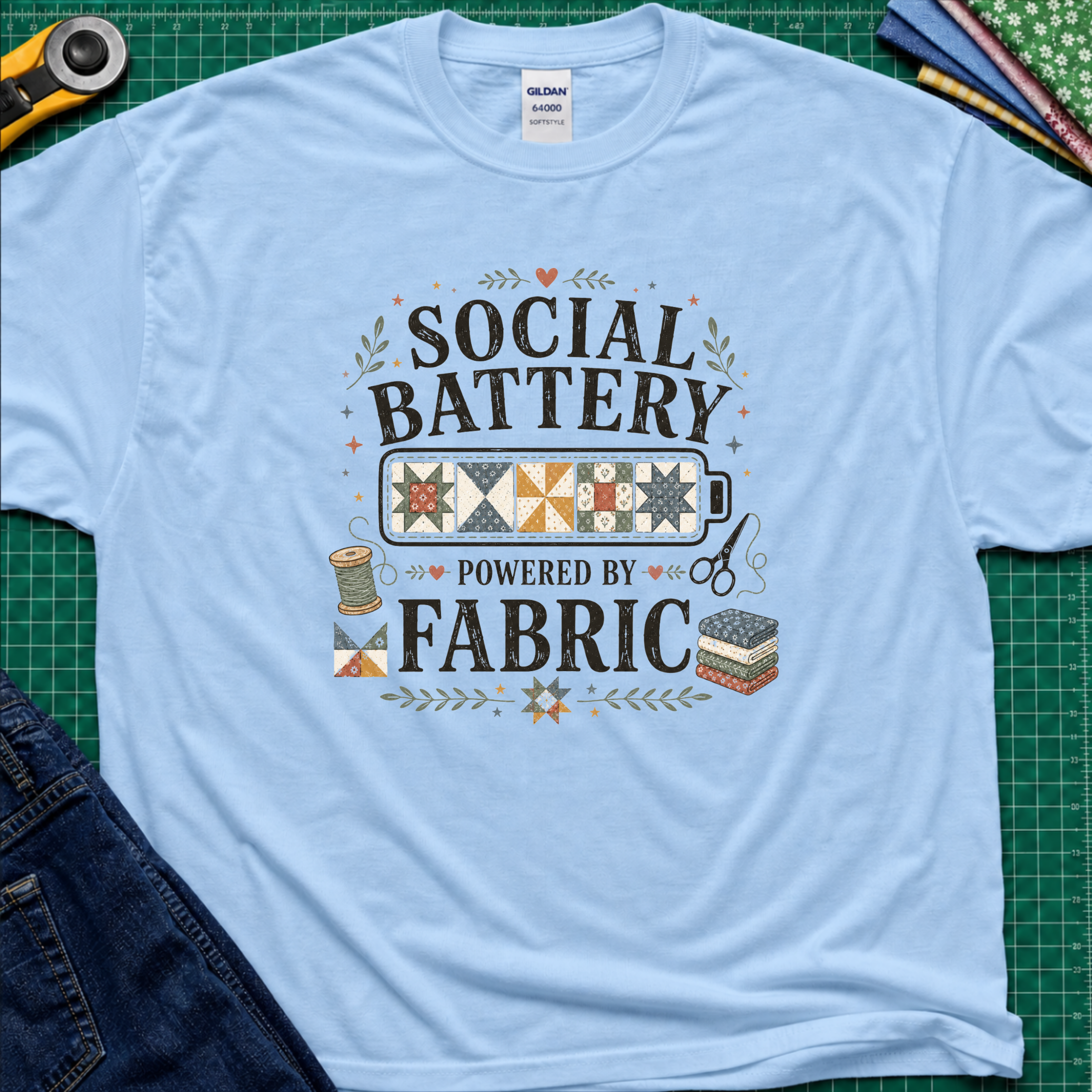 A light blue Gildan t-shirt with a 'Social Battery Powered by Fabric' graphic, laid flat on a green cutting mat with sewing props like a rotary cutter, scissors, and fabric rolls.