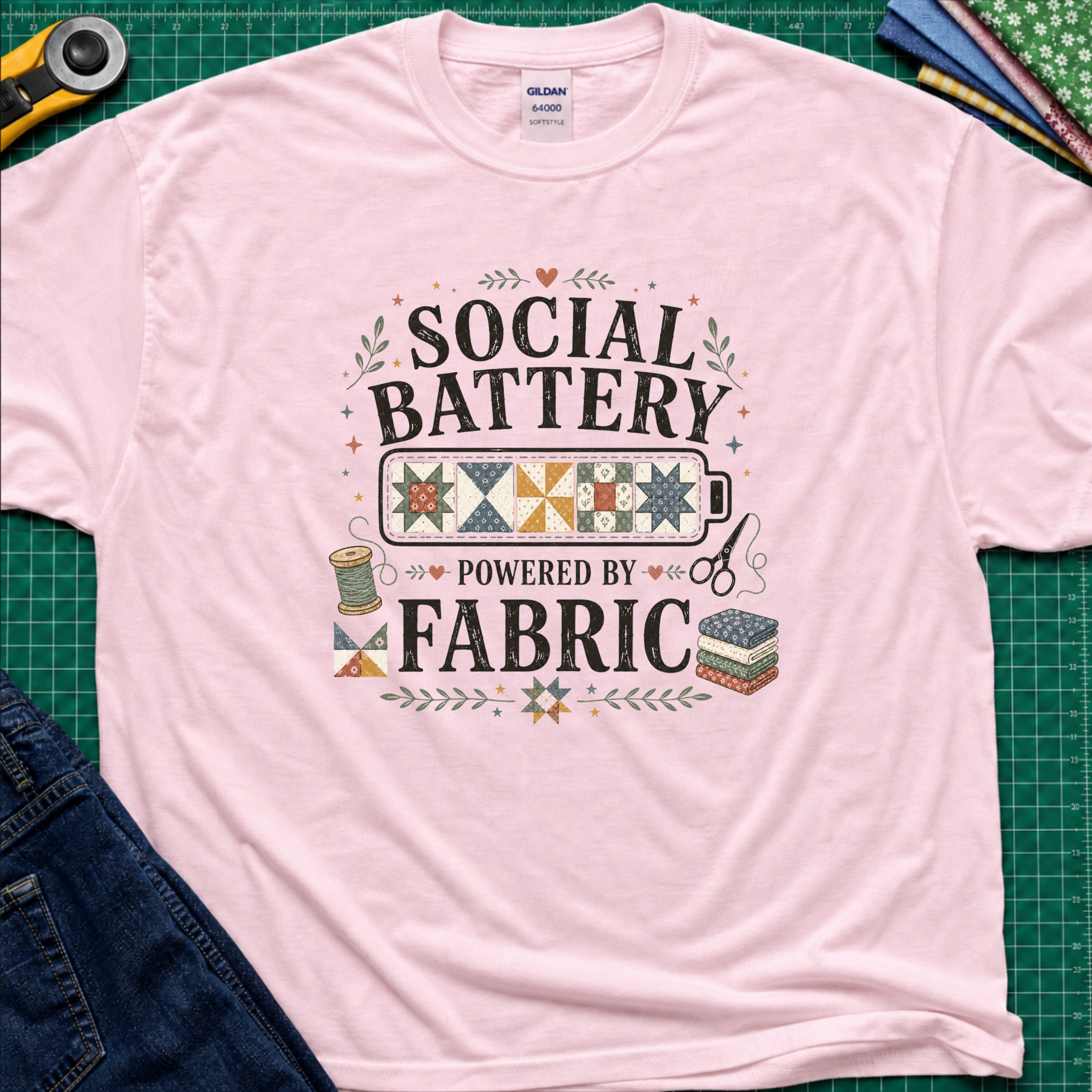 A light pink Gildan t-shirt with a 'Social Battery Powered by Fabric' graphic, laid flat on a green cutting mat with sewing props like a rotary cutter, scissors, and fabric rolls.