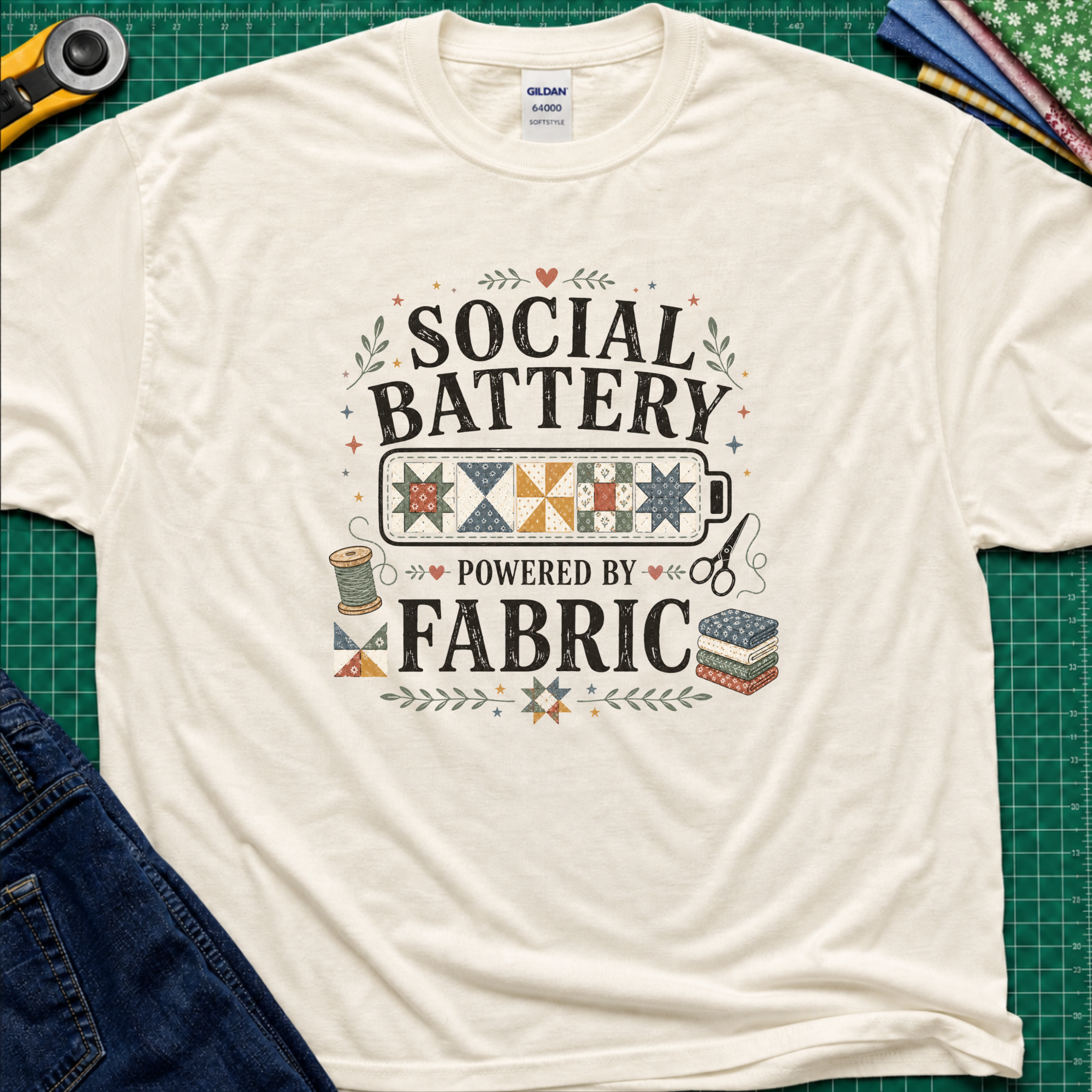 A light cream Gildan t-shirt with a 'Social Battery Powered by Fabric' graphic, laid flat on a green cutting mat with sewing props like a rotary cutter, scissors, and fabric rolls.