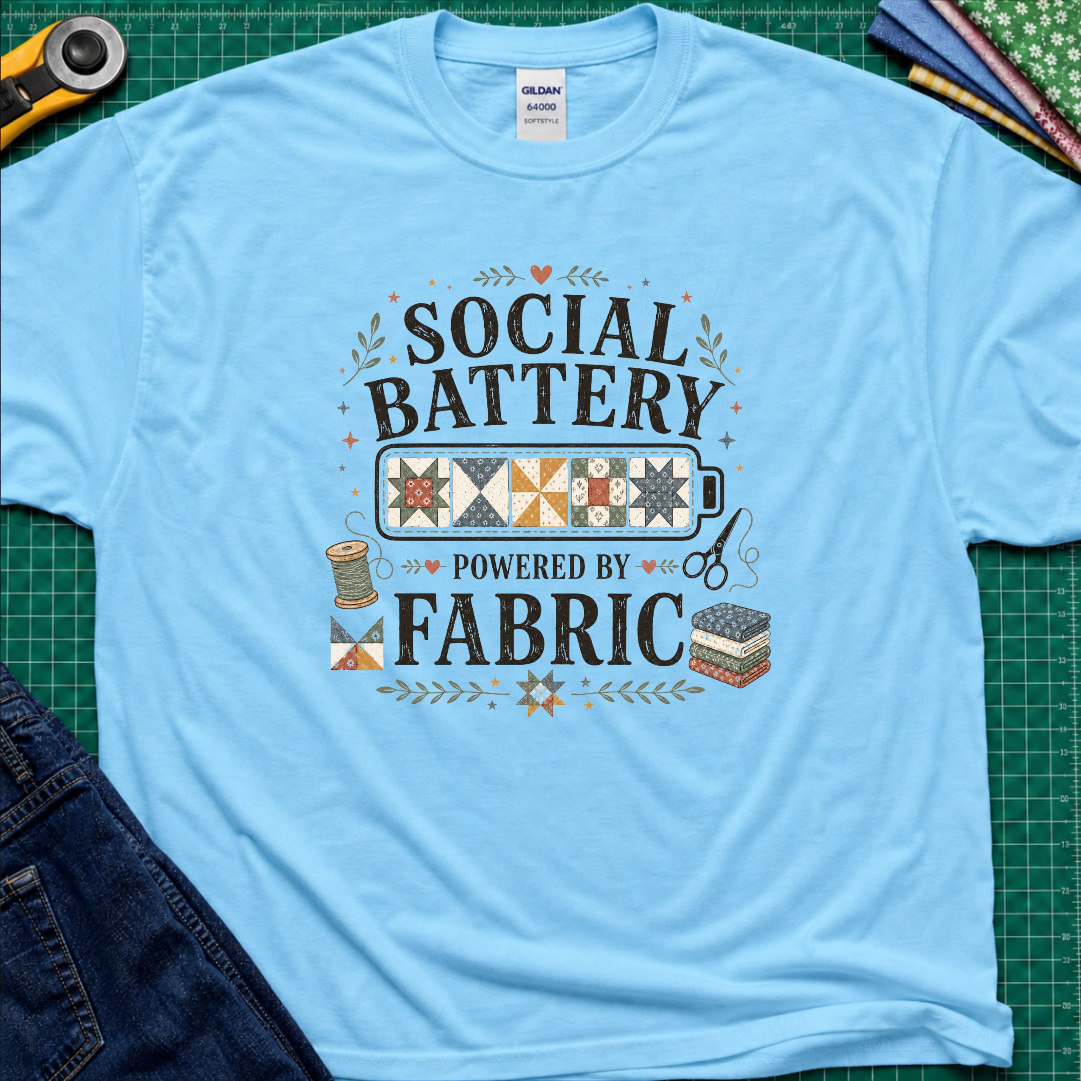 A sky blue Gildan t-shirt with a 'Social Battery Powered by Fabric' graphic, laid flat on a green cutting mat with sewing props like a rotary cutter, scissors, and fabric rolls.
