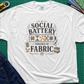 A white Gildan t-shirt with a 'Social Battery Powered by Fabric' graphic, laid flat on a green cutting mat with sewing props like a rotary cutter, scissors, and fabric rolls.