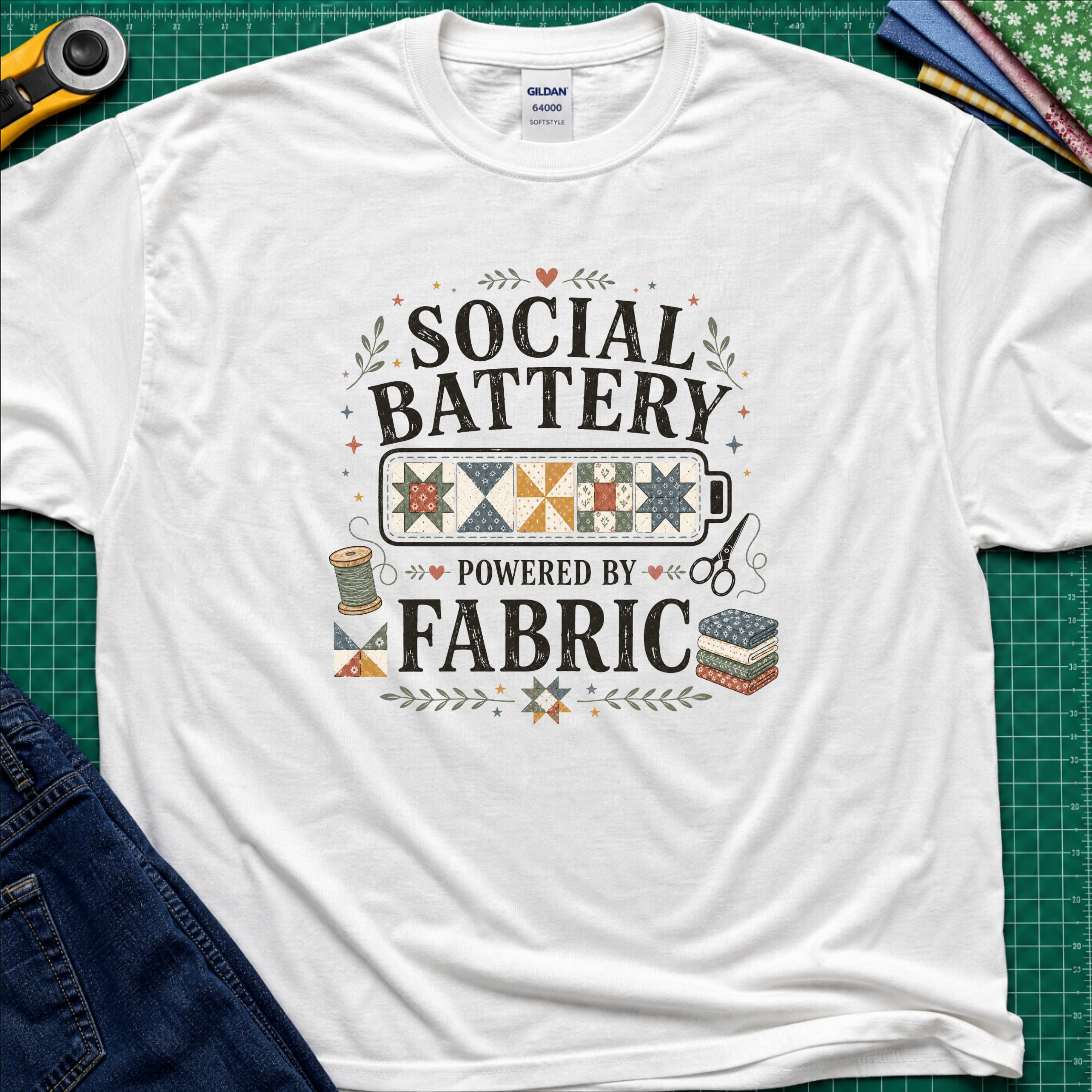 A white Gildan t-shirt with a 'Social Battery Powered by Fabric' graphic, laid flat on a green cutting mat with sewing props like a rotary cutter, scissors, and fabric rolls.