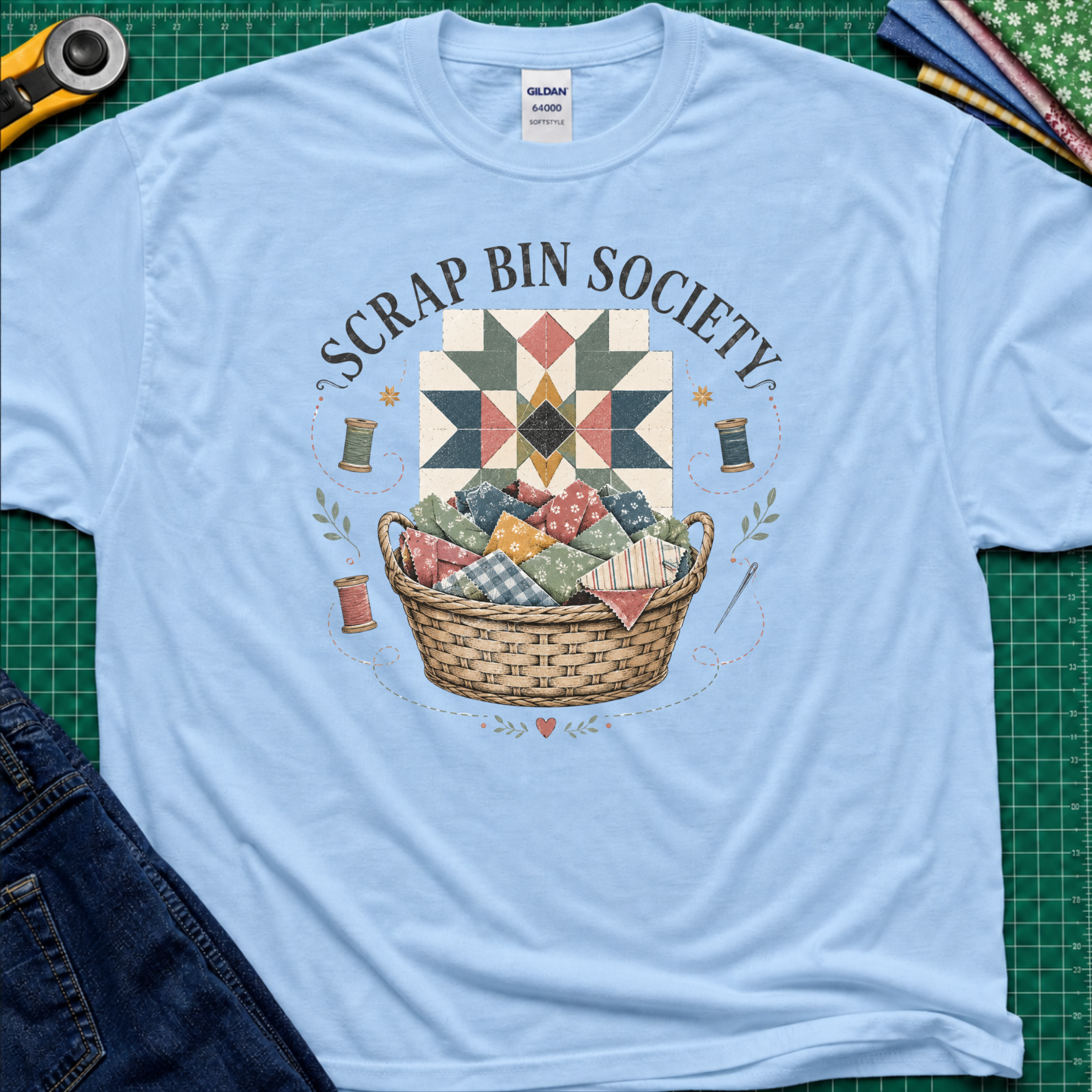 A light blue Gildan t-shirt with a 'Scrap Bin Society' quilt basket graphic, styled flat-lay on a green cutting mat with sewing tools and denim jeans.