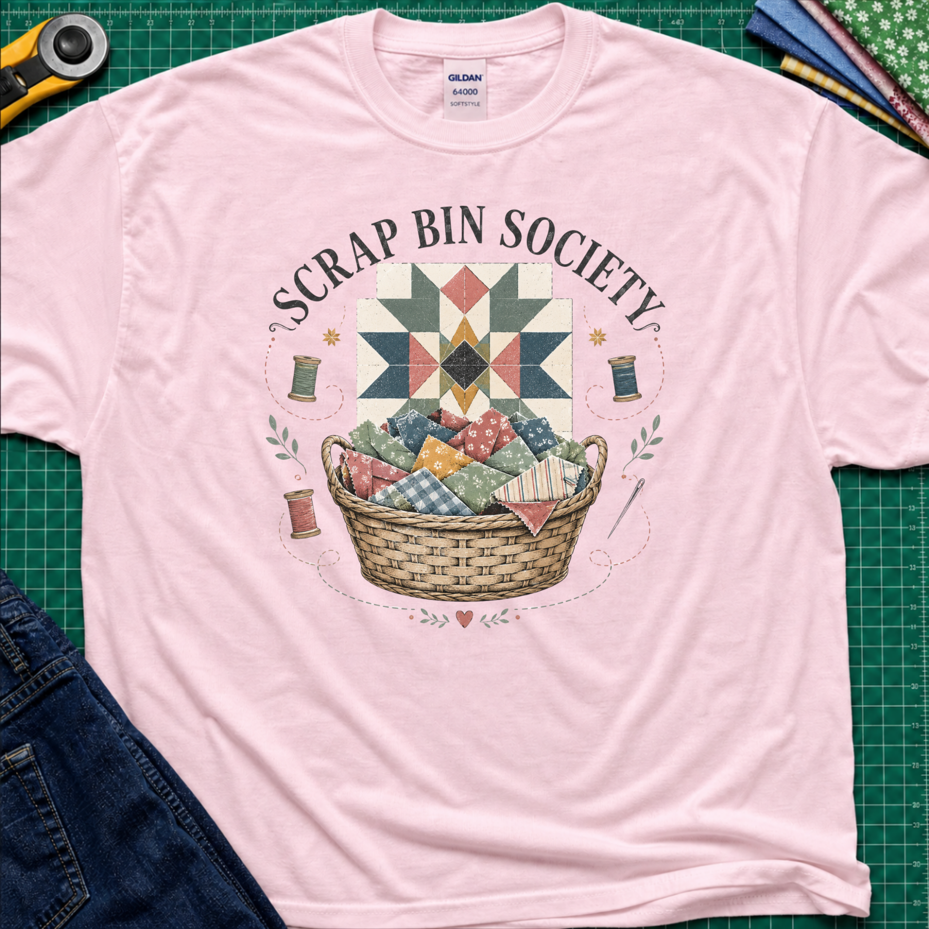 A light pink Gildan t-shirt with a 'Scrap Bin Society' quilt basket graphic, styled flat-lay on a green cutting mat with sewing tools and denim jeans.