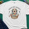 A white Gildan t-shirt with a 'Scrap Bin Society' quilt basket graphic, styled flat-lay on a green cutting mat with sewing tools and denim jeans.
