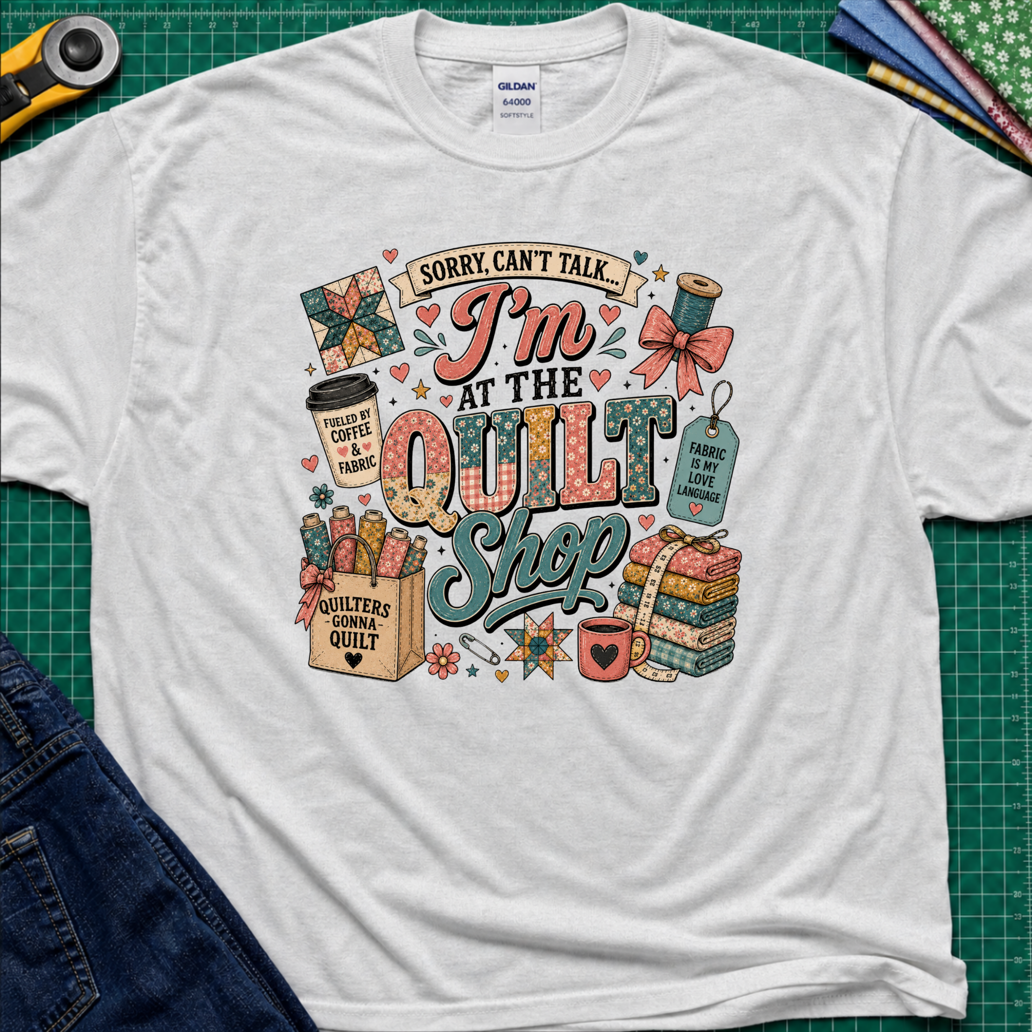A light ice grey Gildan t-shirt with a colorful graphic design that reads 'Sorry, can't talk I'm at the quilt shop' surrounded by quilting-themed illustrations, laid flat on a green cutting mat with sewing props.