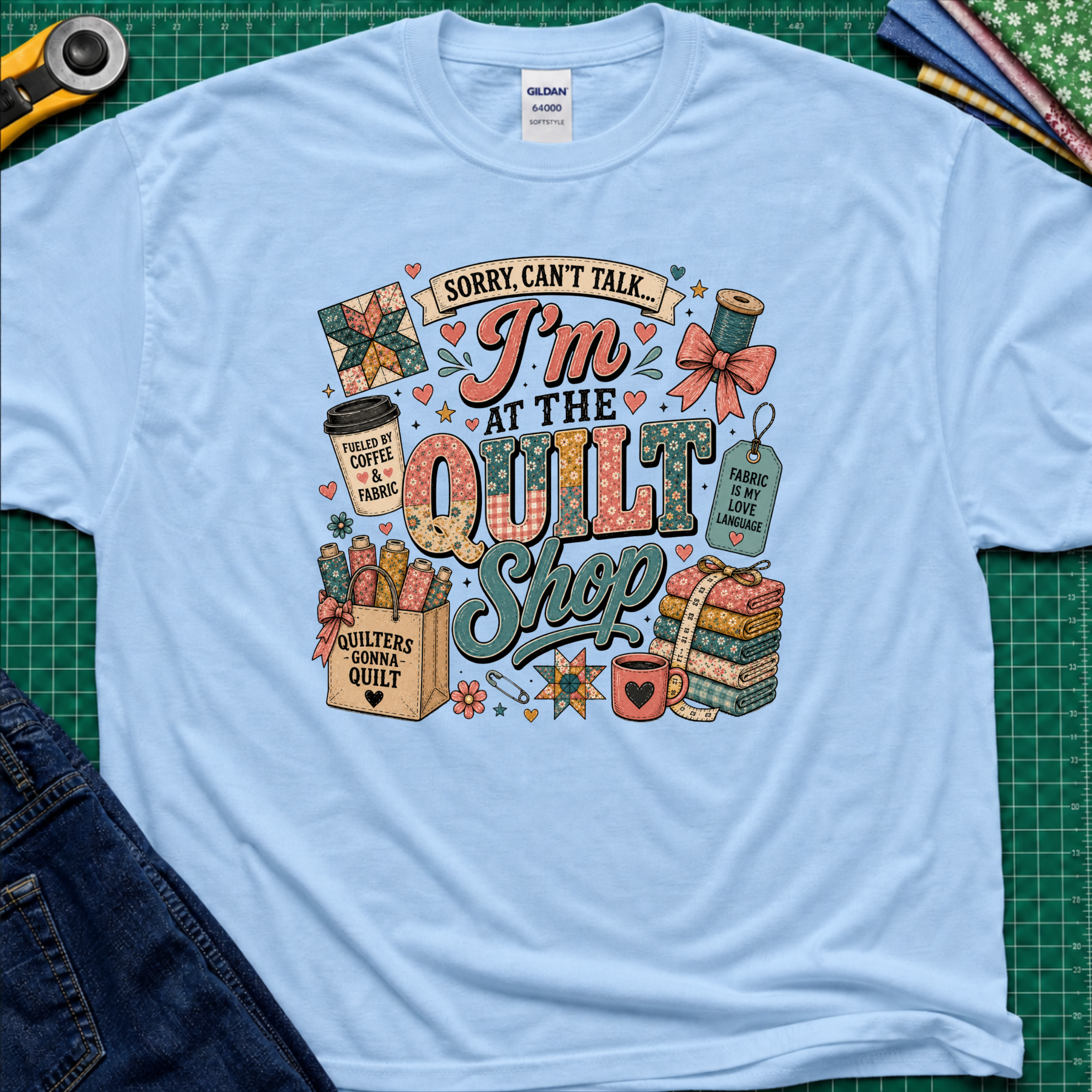 A light blue Gildan t-shirt with a colorful graphic design that reads 'Sorry, can't talk I'm at the quilt shop' surrounded by quilting-themed illustrations, laid flat on a green cutting mat with sewing props.