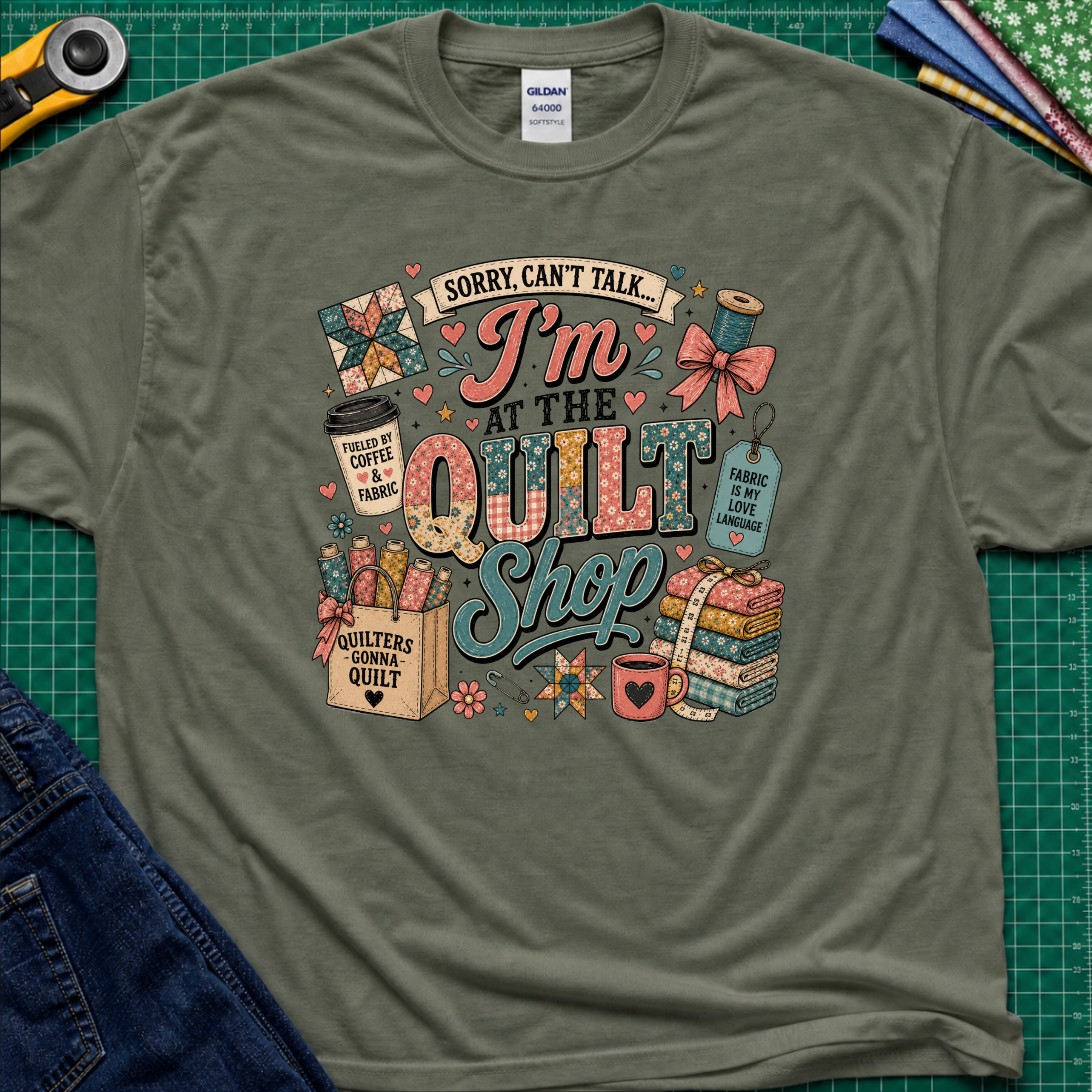 A military green Gildan t-shirt with a colorful graphic design that reads 'Sorry, can't talk I'm at the quilt shop' surrounded by quilting-themed illustrations, laid flat on a green cutting mat with sewing props.