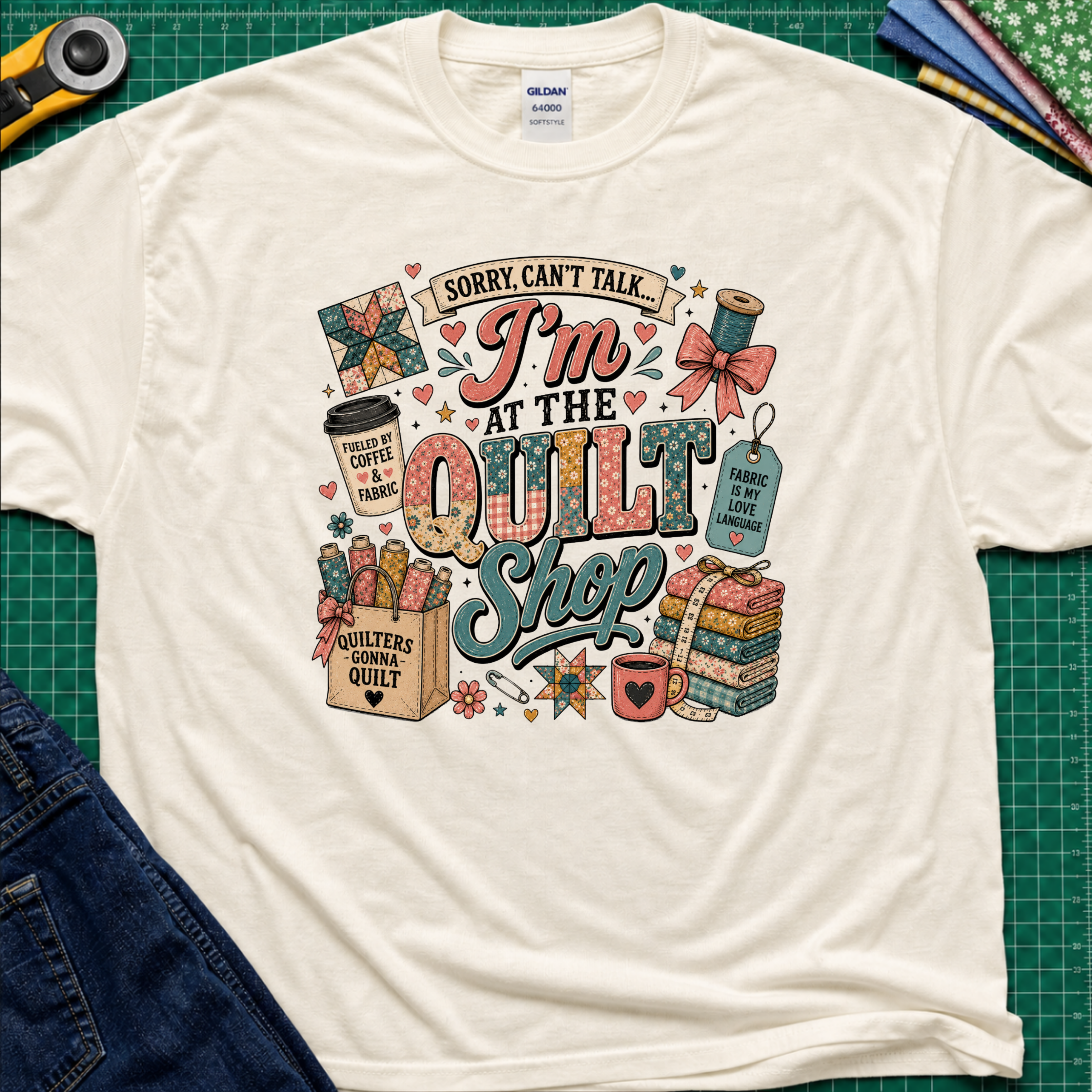 A light cream Gildan t-shirt with a colorful graphic design that reads 'Sorry, can't talk I'm at the quilt shop' surrounded by quilting-themed illustrations, laid flat on a green cutting mat with sewing props.