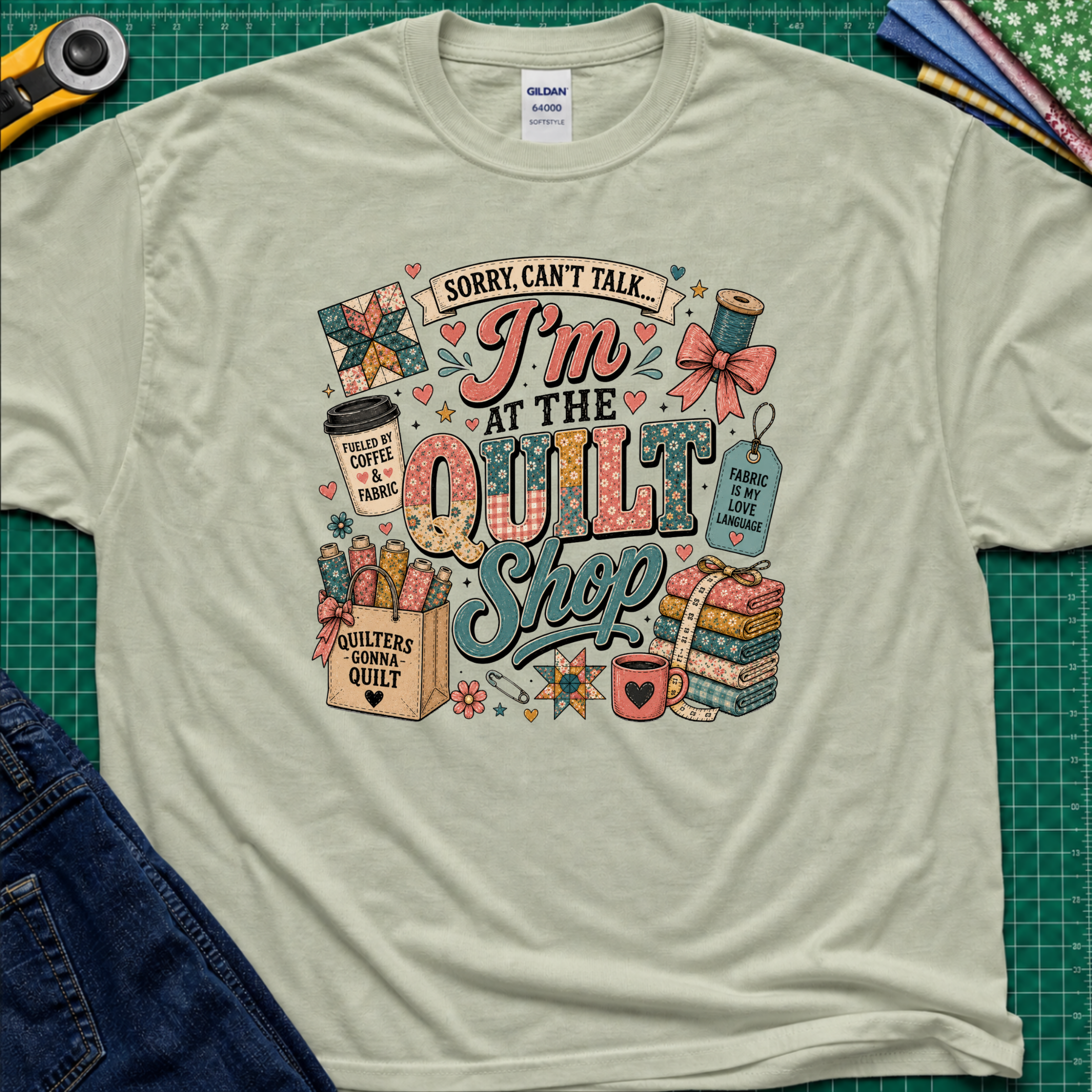 A light sage green Gildan t-shirt with a colorful graphic design that reads 'Sorry, can't talk I'm at the quilt shop' surrounded by quilting-themed illustrations, laid flat on a green cutting mat with sewing props.