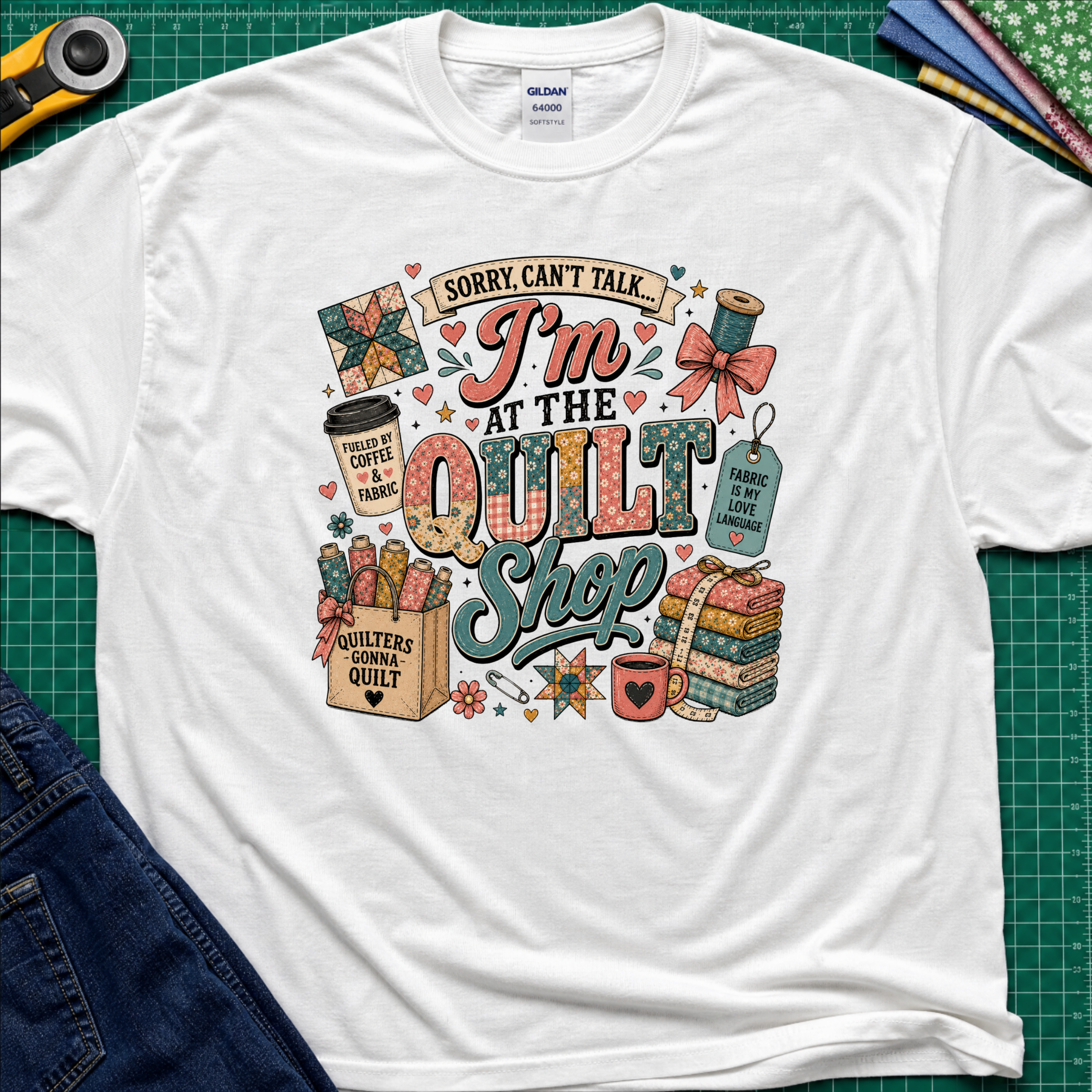 A white Gildan t-shirt with a colorful graphic design that reads 'Sorry, can't talk I'm at the quilt shop' surrounded by quilting-themed illustrations, laid flat on a green cutting mat with sewing props.