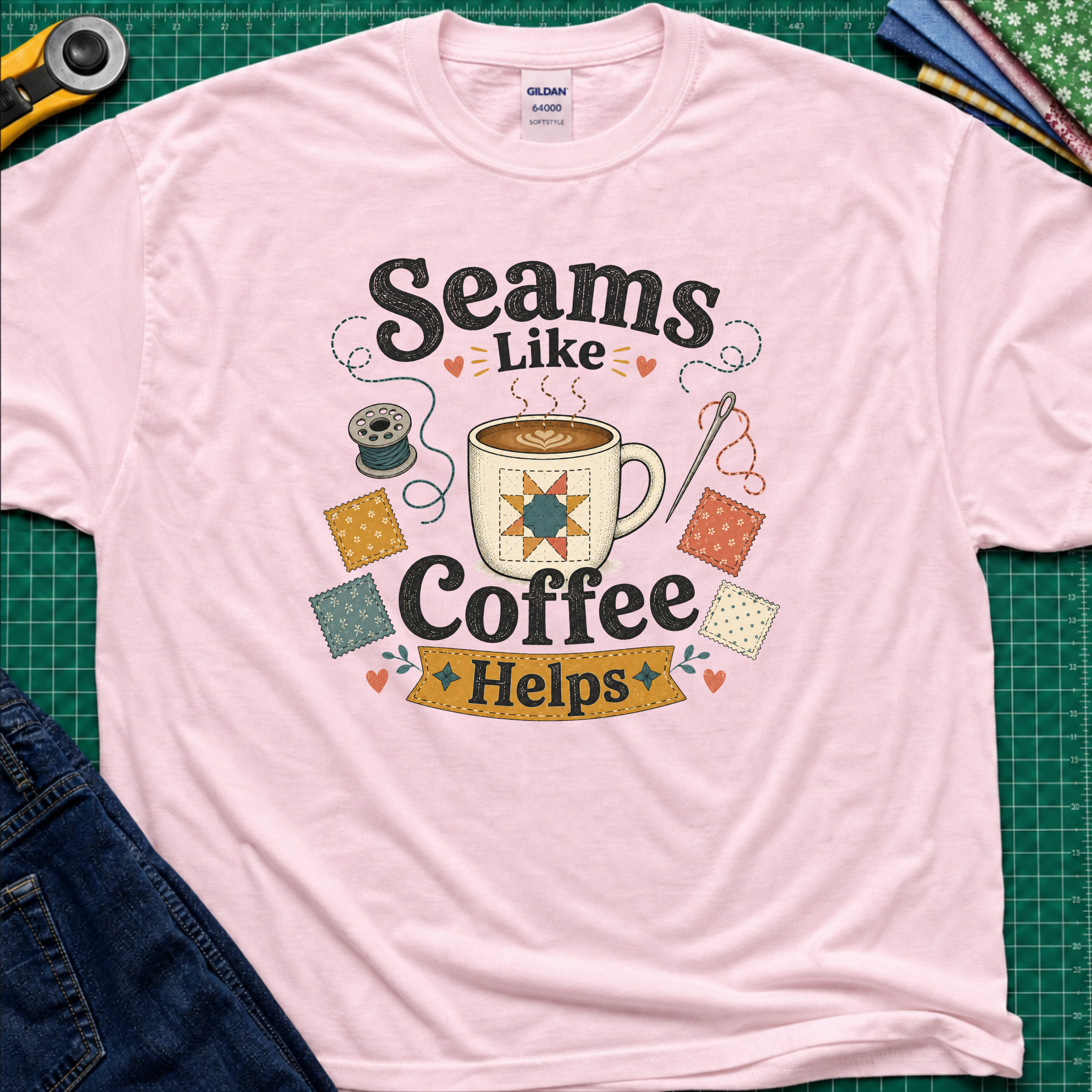 A light pink Gildan t-shirt with a colorful graphic that reads 'Seams Like Coffee Helps' with a coffee mug, fabric swatches, needle and bobbin.