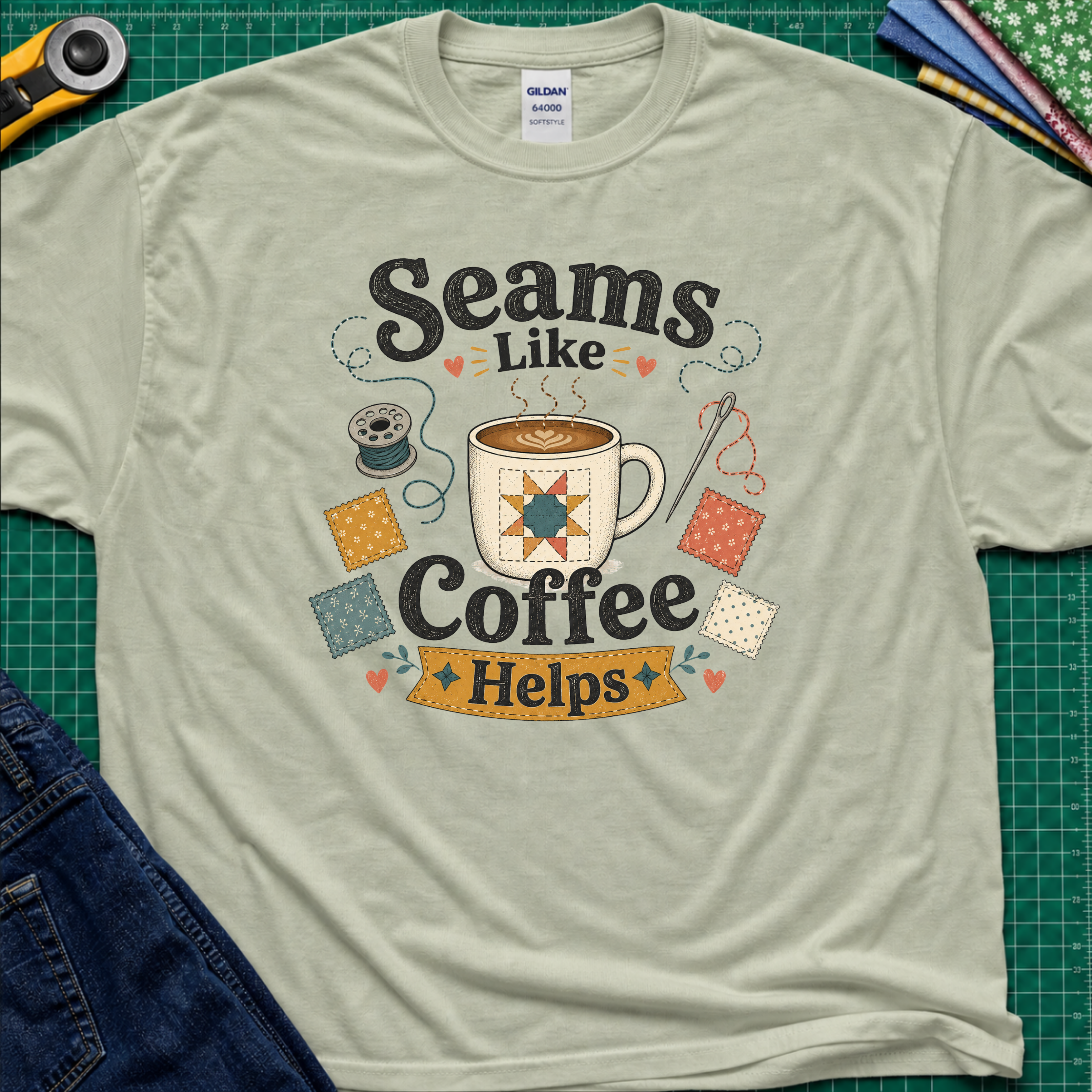 A light sage green Gildan t-shirt with a colorful graphic that reads 'Seams Like Coffee Helps' with a coffee mug, fabric swatches, needle and bobbin.