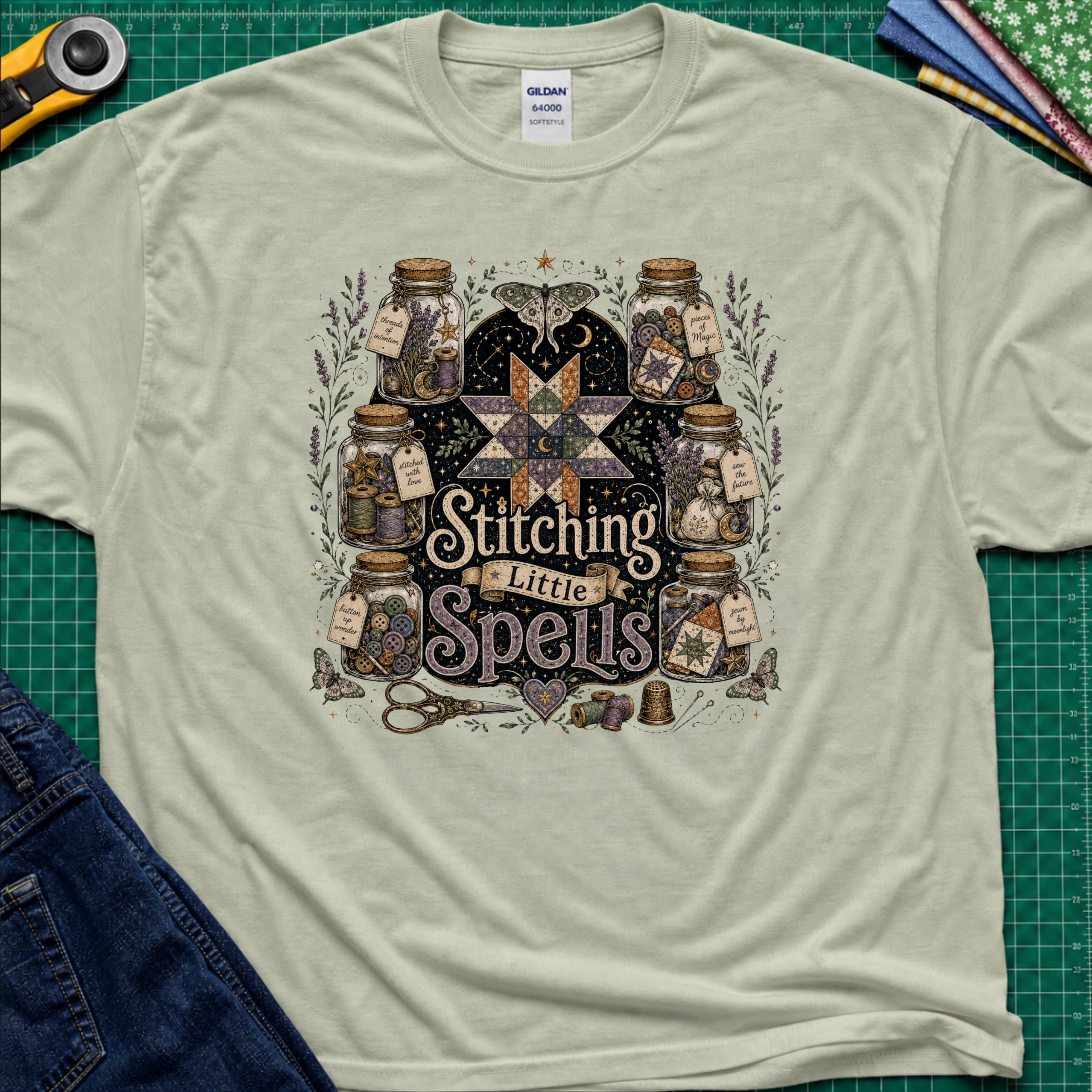 A light sage green Gildan t-shirt laid flat on a green cutting mat, featuring a detailed graphic of vintage glass jars, quills, scissors, and a star with the text 'Stitching Little Spells'.