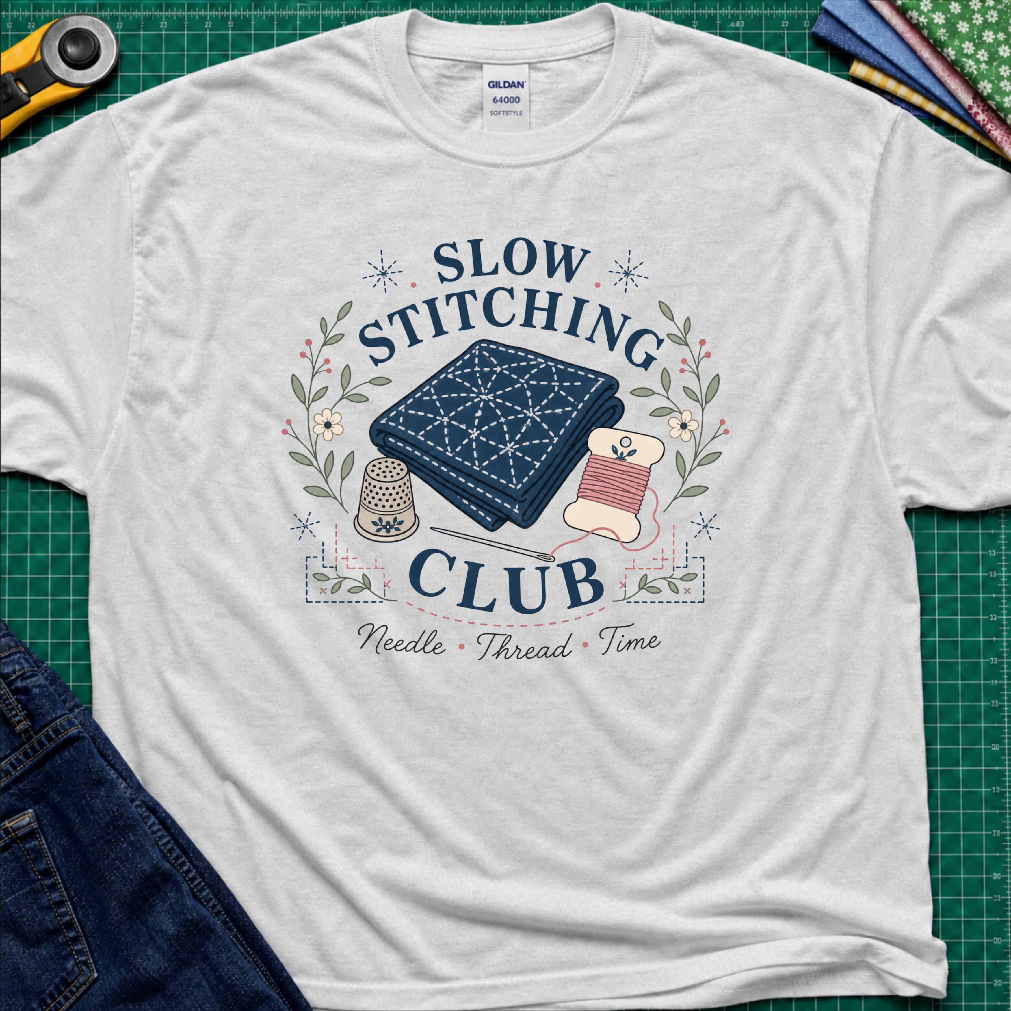 A light grey Gildan t-shirt featuring a 'Slow Stitching Club' graphic with navy sashiko stitched fabric, thimble, thread, with floral accents. The shirt lays flat on a green cutting mat with a rotary cutter, fabric stash and blue jeans partially visible in the corners.