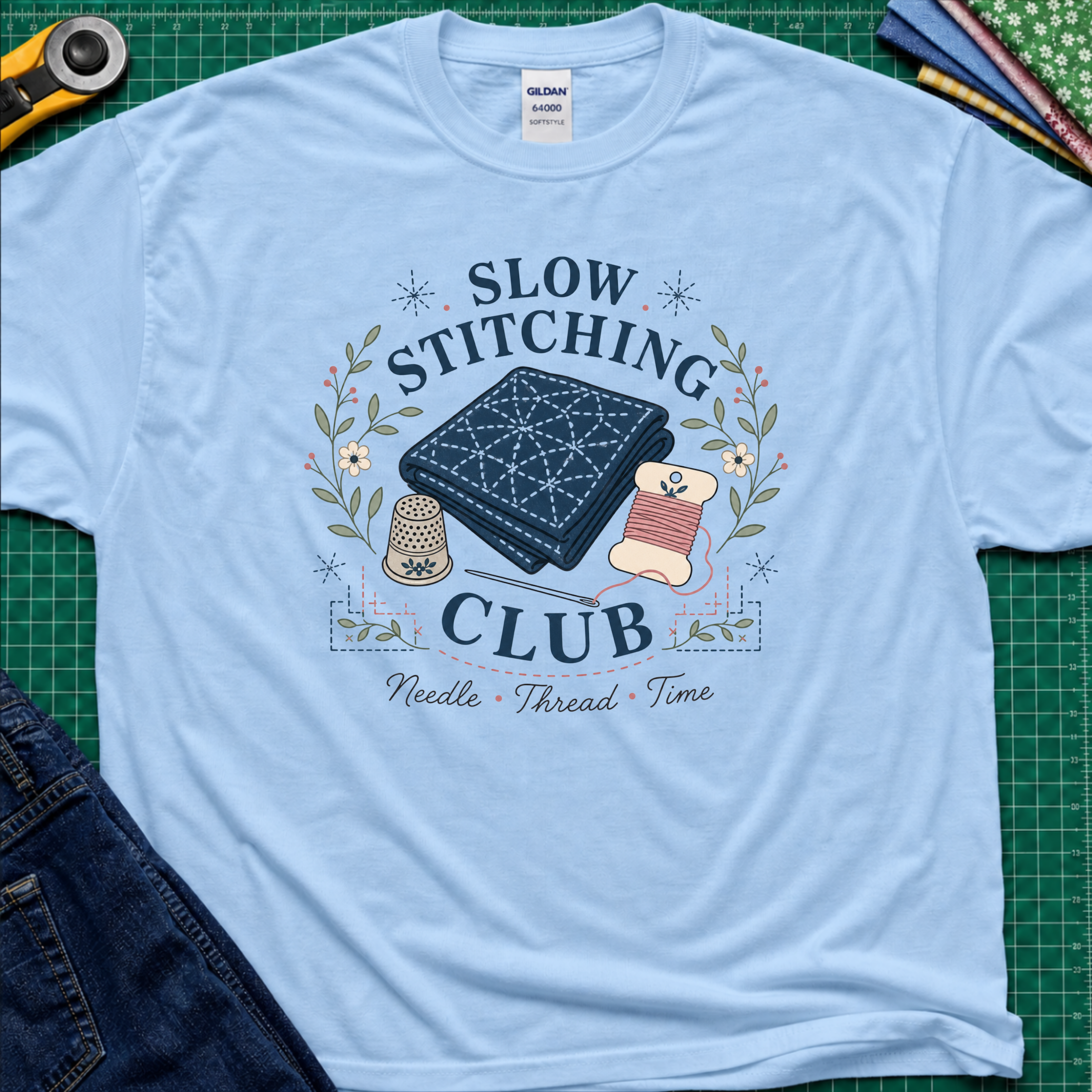 A light blue Gildan t-shirt featuring a 'Slow Stitching Club' graphic with navy sashiko stitched fabric, thimble, thread, with floral accents. The shirt lays flat on a green cutting mat with a rotary cutter, fabric stash and blue jeans partially visible in the corners.
