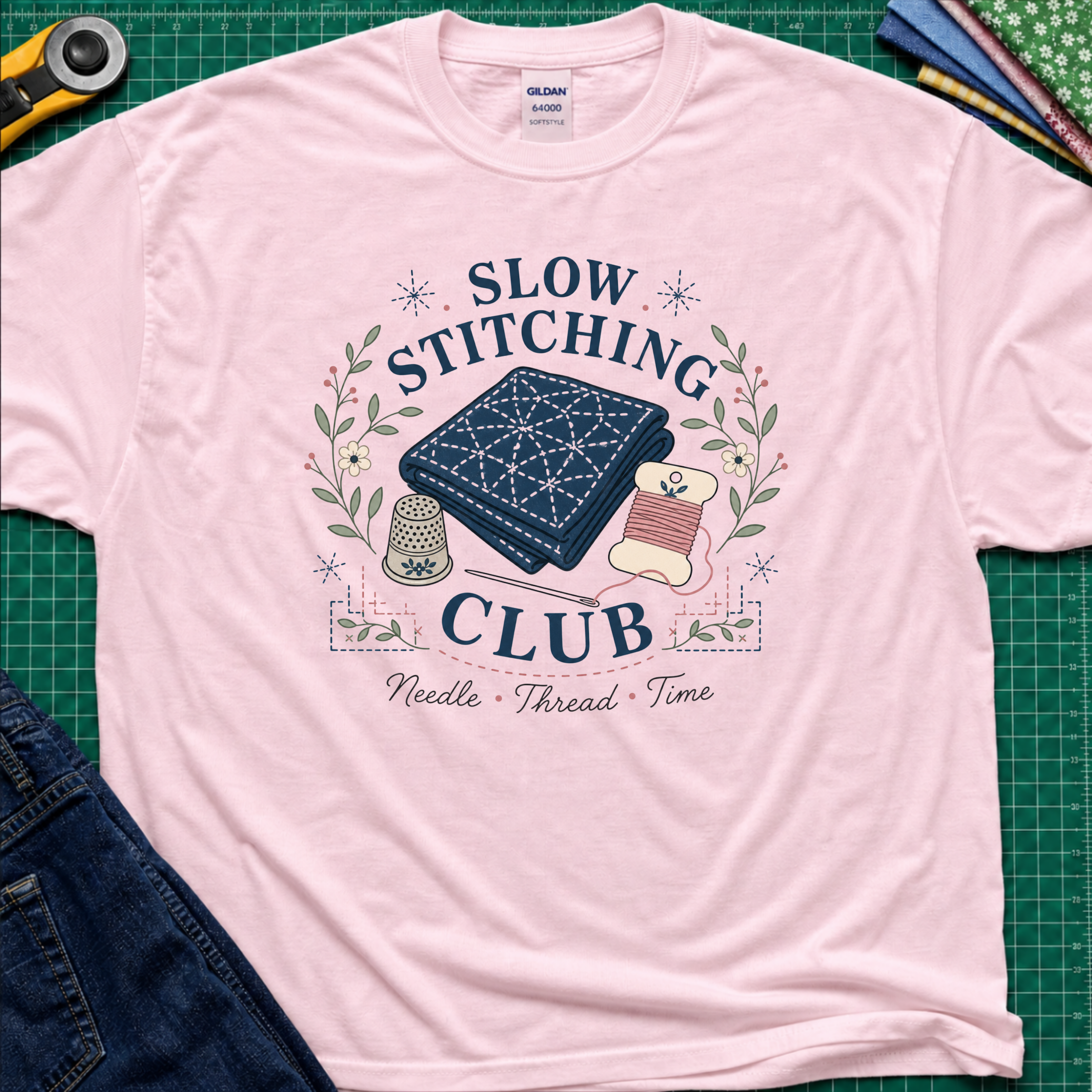 A light pink Gildan t-shirt featuring a 'Slow Stitching Club' graphic with navy sashiko stitched fabric, thimble, thread, with floral accents. The shirt lays flat on a green cutting mat with a rotary cutter, fabric stash and blue jeans partially visible in the corners.