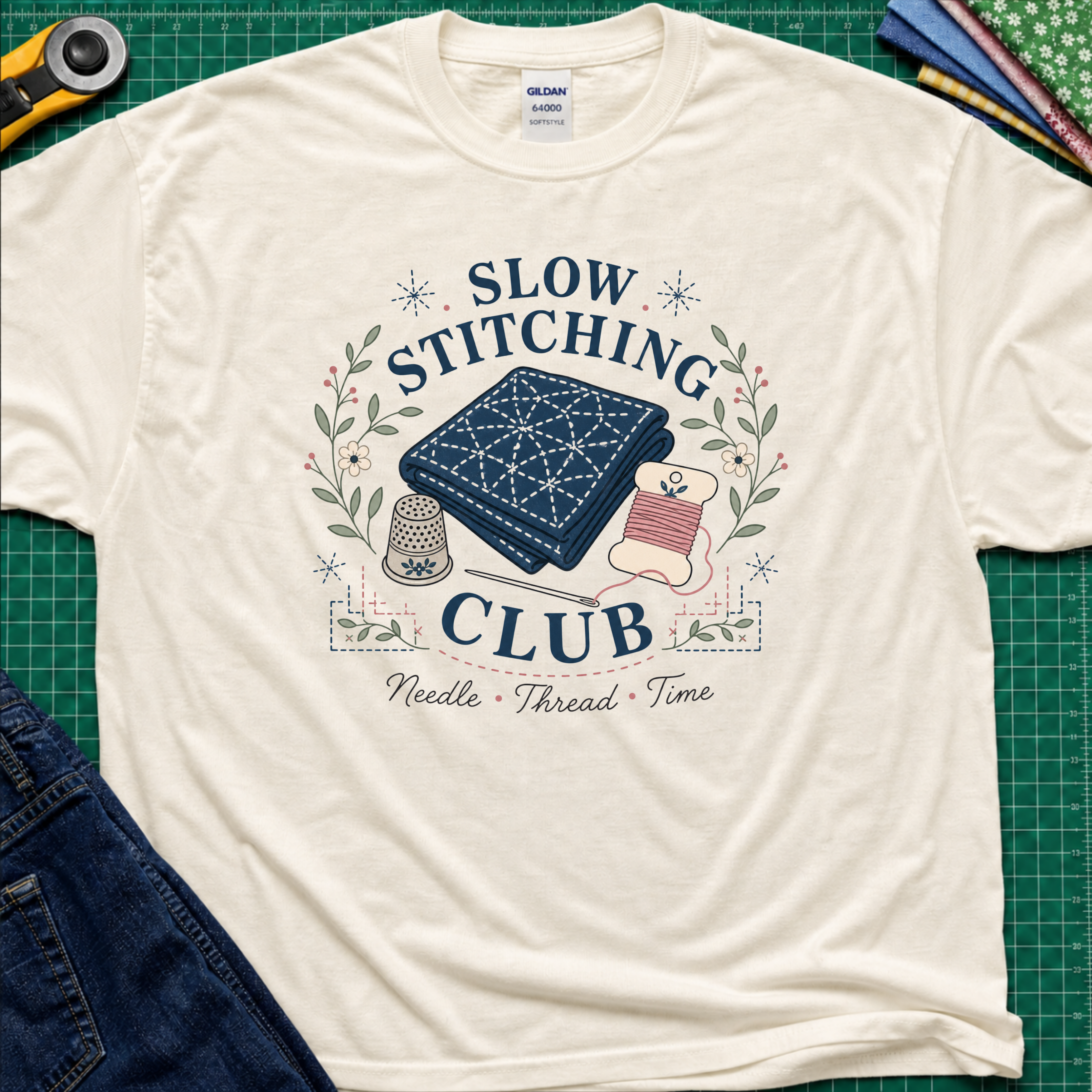 A light cream Gildan t-shirt featuring a 'Slow Stitching Club' graphic with navy sashiko stitched fabric, thimble, thread, with floral accents. The shirt lays flat on a green cutting mat with a rotary cutter, fabric stash and blue jeans partially visible in the corners.