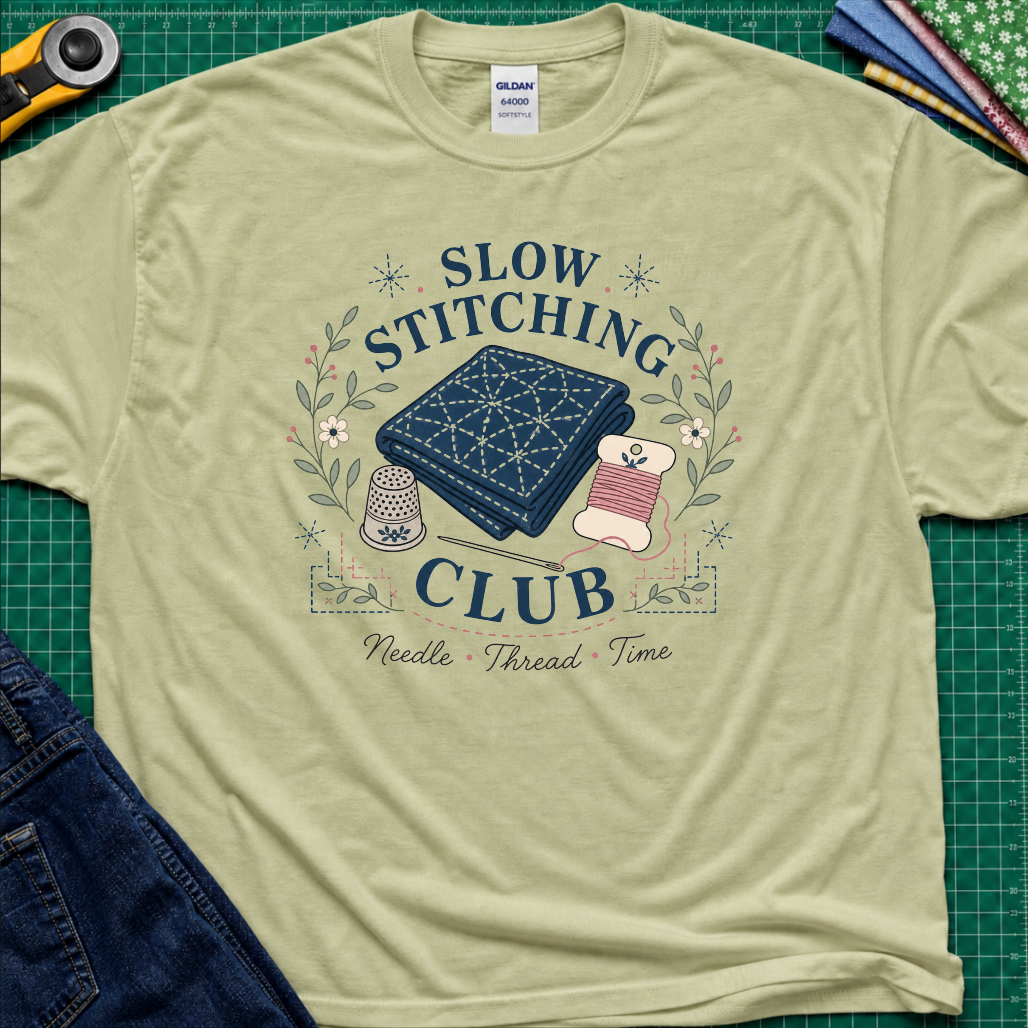 A light pistachio green Gildan t-shirt featuring a 'Slow Stitching Club' graphic with navy sashiko stitched fabric, thimble, thread, with floral accents. The shirt lays flat on a green cutting mat with a rotary cutter, fabric stash and blue jeans partially visible in the corners.