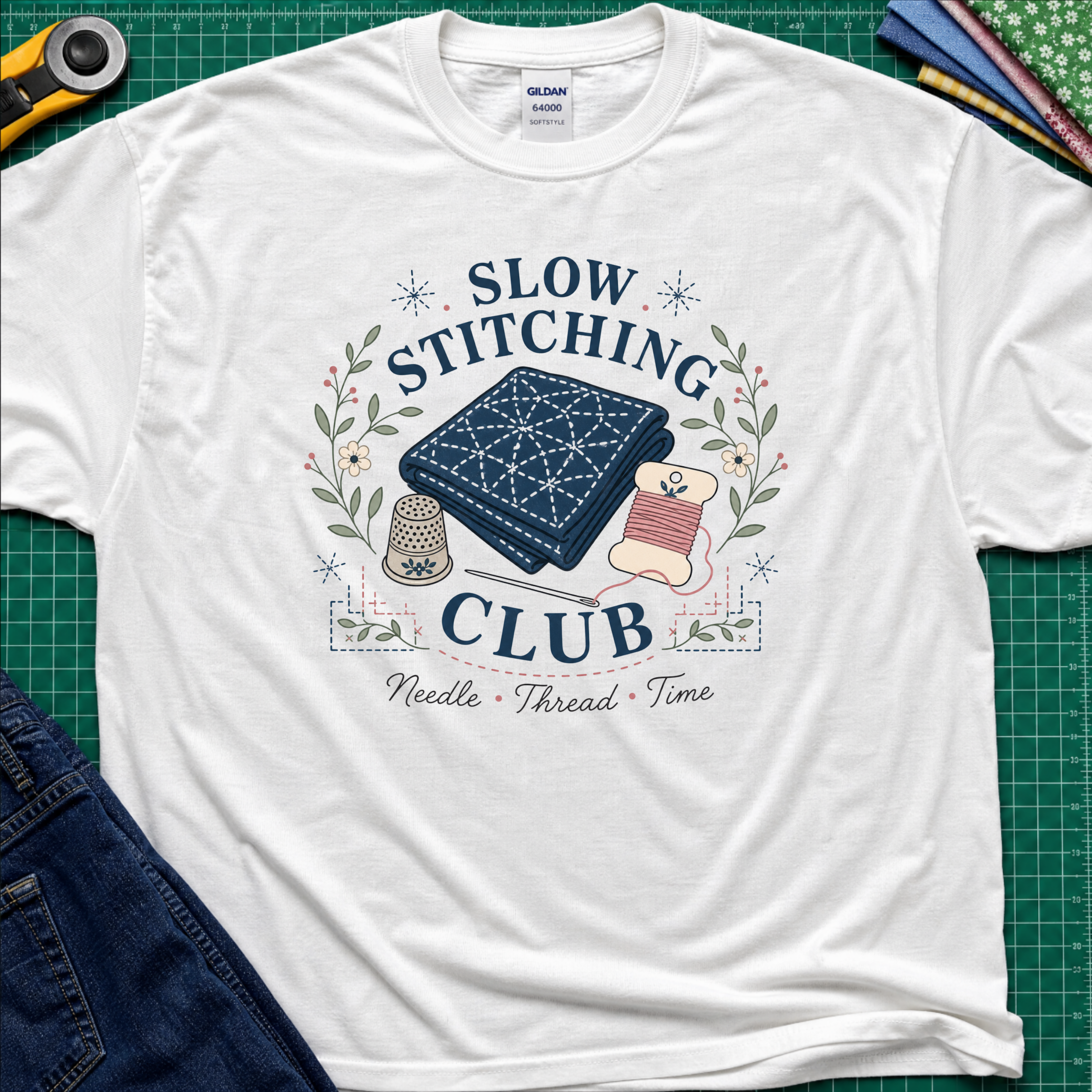 A white Gildan t-shirt featuring a 'Slow Stitching Club' graphic with navy sashiko stitched fabric, thimble, thread, with floral accents. The shirt lays flat on a green cutting mat with a rotary cutter, fabric stash and blue jeans partially visible in the corners.