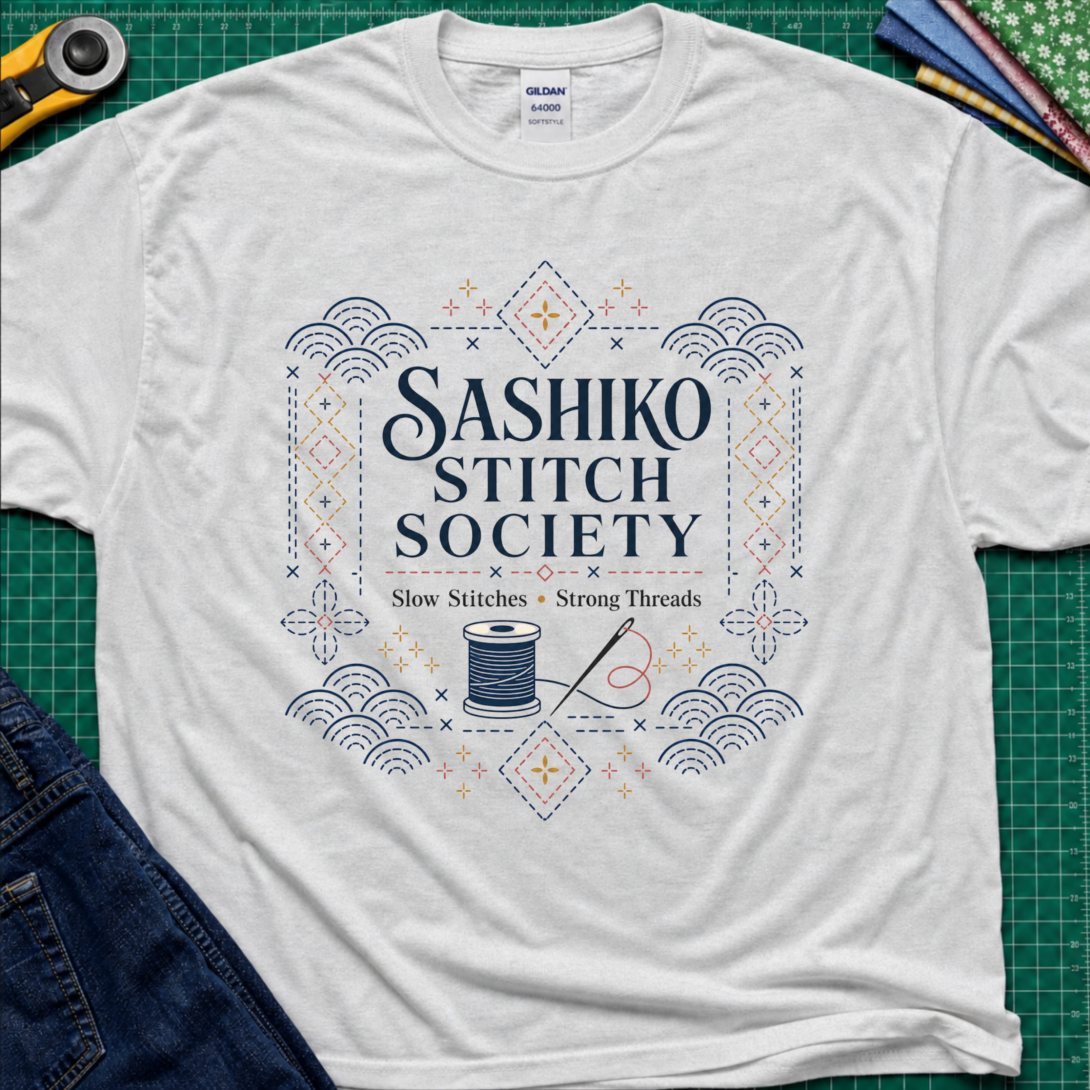 A light grey Gildan t-shirt featuring a decorative sashiko style graphic that reads 'SASHIKO STITCH SOCIETY' with a spool of thread and needle illustration. The shirt is laid flat on a green cutting mat with a rotary cutter, fabric stack and blue jeans partially visible in the corners.