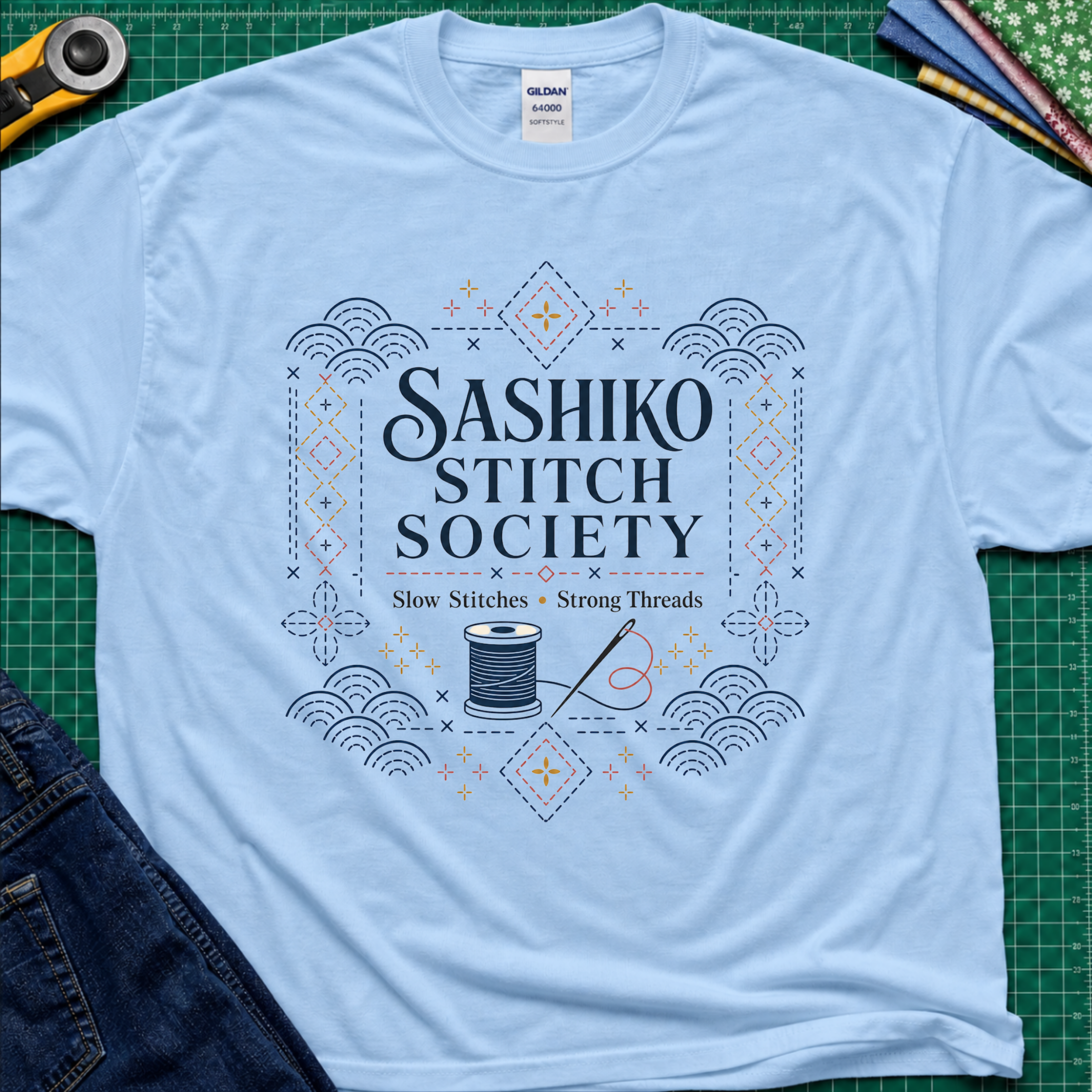 A light blue Gildan t-shirt featuring a decorative sashiko style graphic that reads 'SASHIKO STITCH SOCIETY' with a spool of thread and needle illustration. The shirt is laid flat on a green cutting mat with a rotary cutter, fabric stack and blue jeans partially visible in the corners.