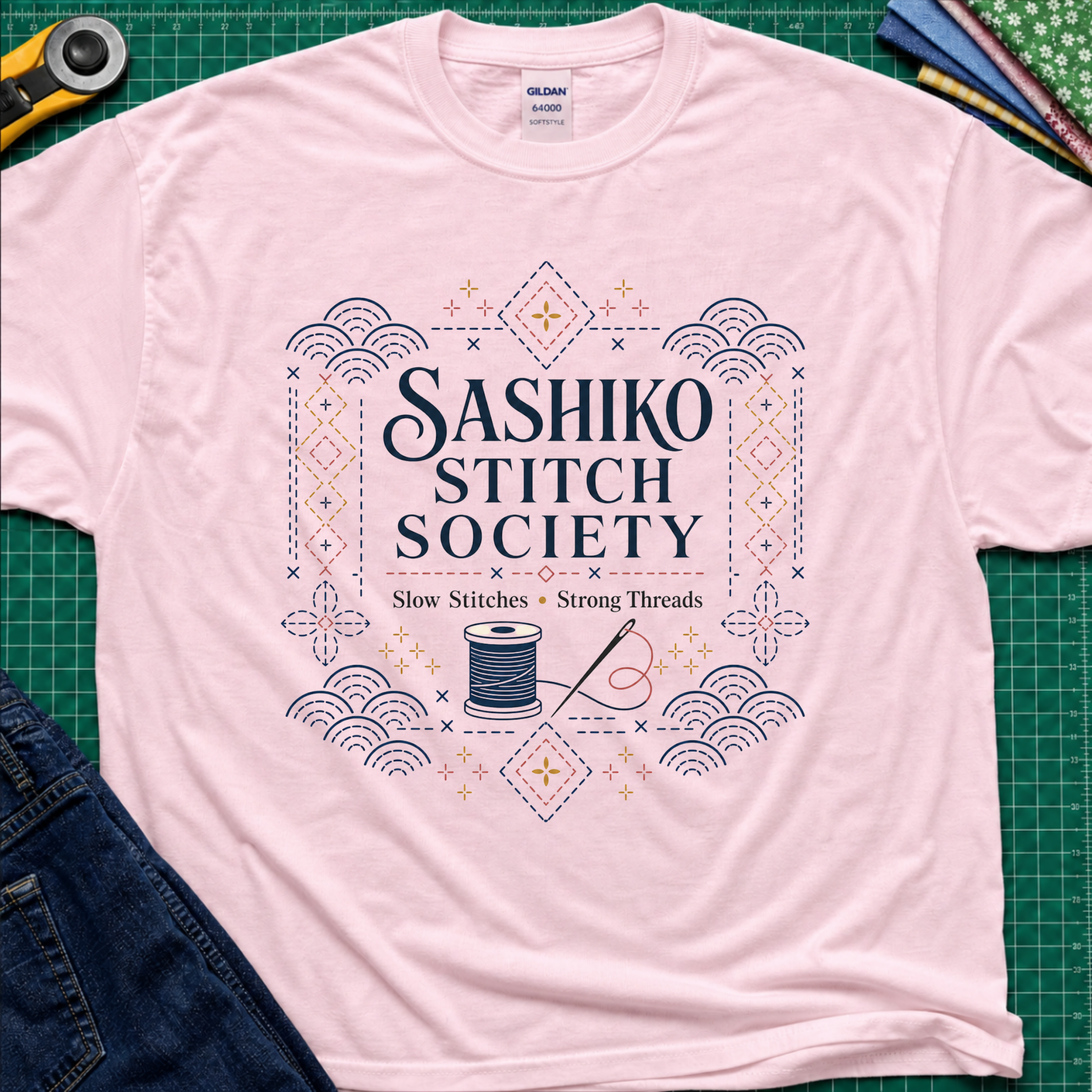 A light pink Gildan t-shirt featuring a decorative sashiko style graphic that reads 'SASHIKO STITCH SOCIETY' with a spool of thread and needle illustration. The shirt is laid flat on a green cutting mat with a rotary cutter, fabric stack and blue jeans partially visible in the corners.
