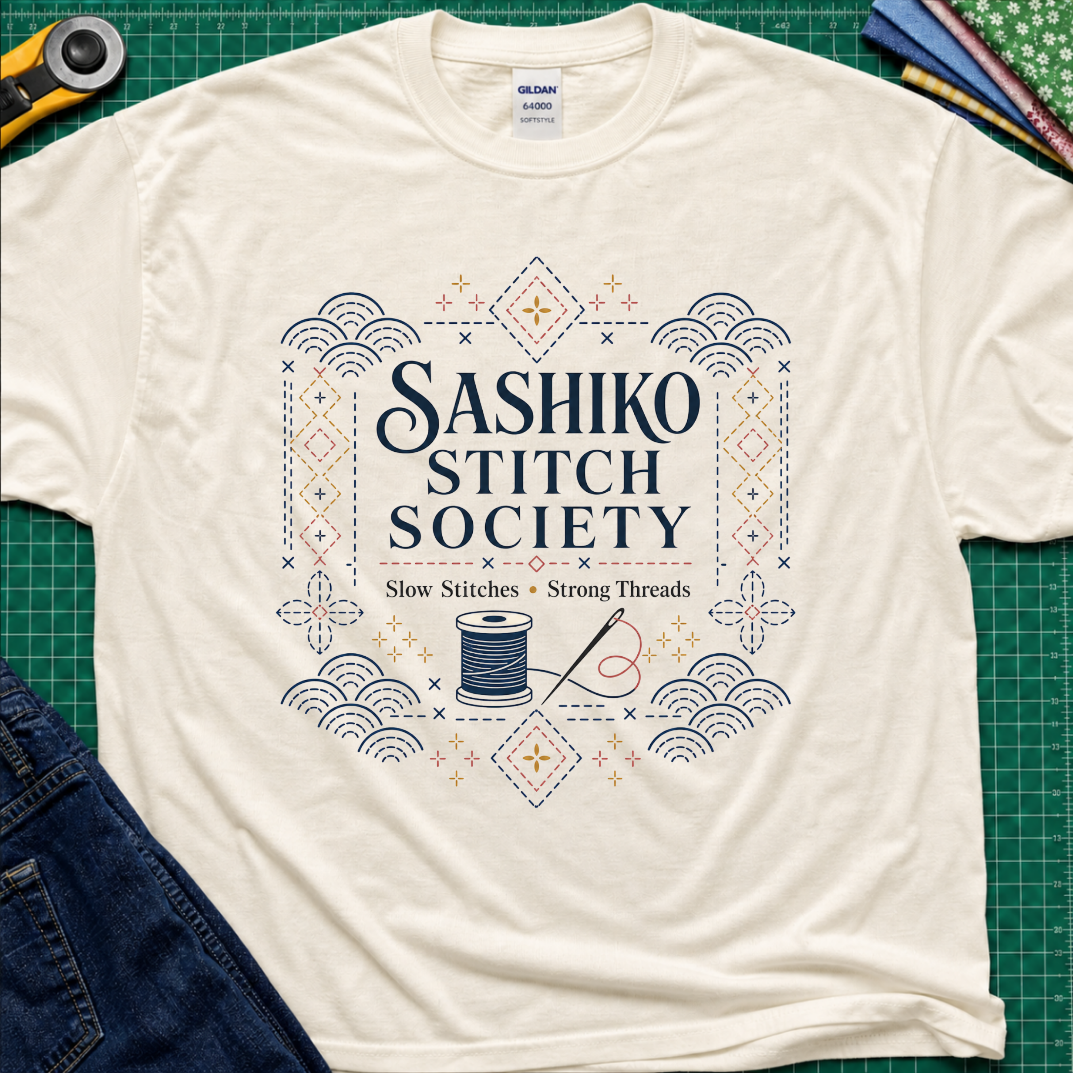 A light cream Gildan t-shirt featuring a decorative sashiko style graphic that reads 'SASHIKO STITCH SOCIETY' with a spool of thread and needle illustration. The shirt is laid flat on a green cutting mat with a rotary cutter, fabric stack and blue jeans partially visible in the corners.