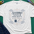 A white Gildan t-shirt featuring a decorative sashiko style graphic that reads 'SASHIKO STITCH SOCIETY' with a spool of thread and needle illustration. The shirt is laid flat on a green cutting mat with a rotary cutter, fabric stack and blue jeans partially visible in the corners.