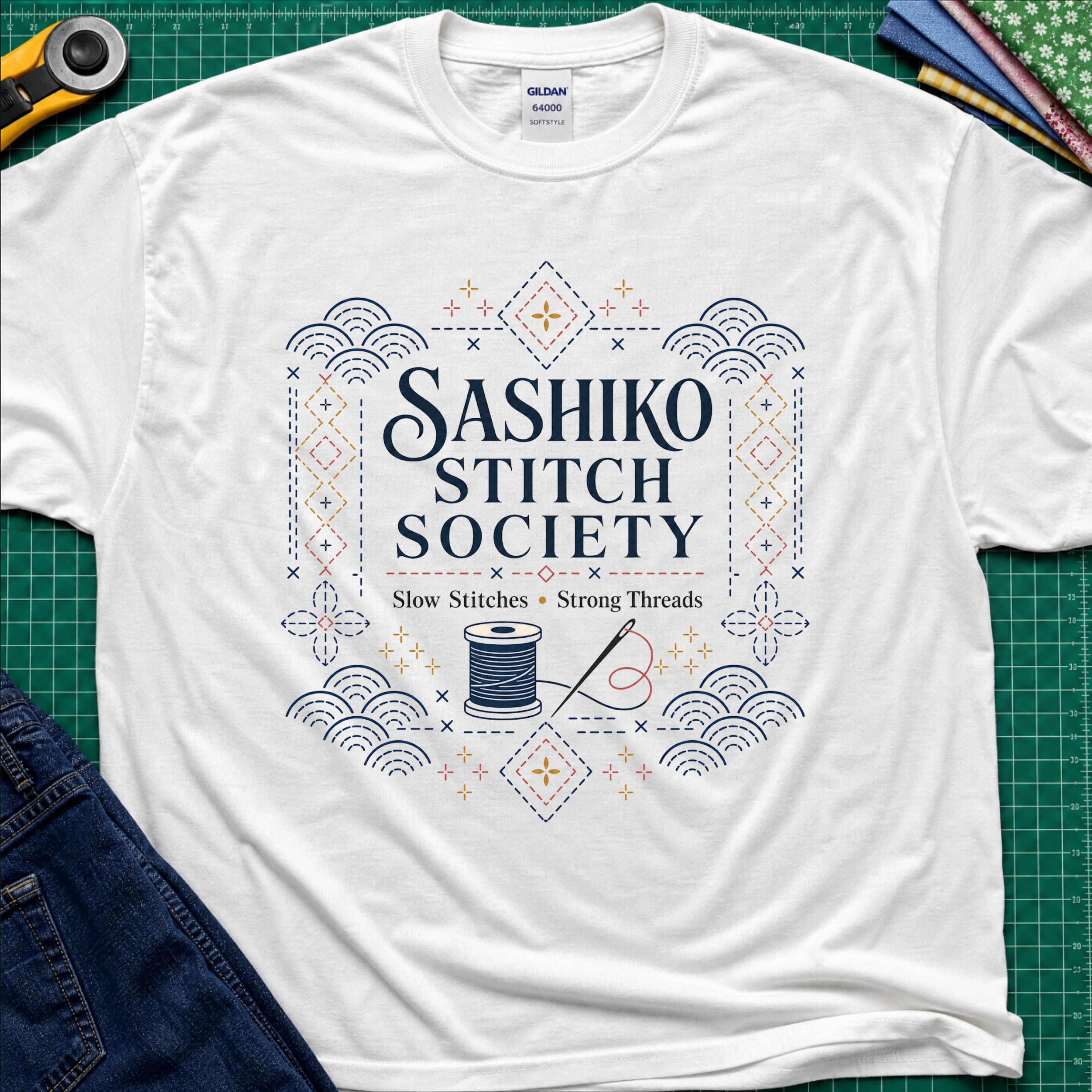 A white Gildan t-shirt featuring a decorative sashiko style graphic that reads 'SASHIKO STITCH SOCIETY' with a spool of thread and needle illustration. The shirt is laid flat on a green cutting mat with a rotary cutter, fabric stack and blue jeans partially visible in the corners.