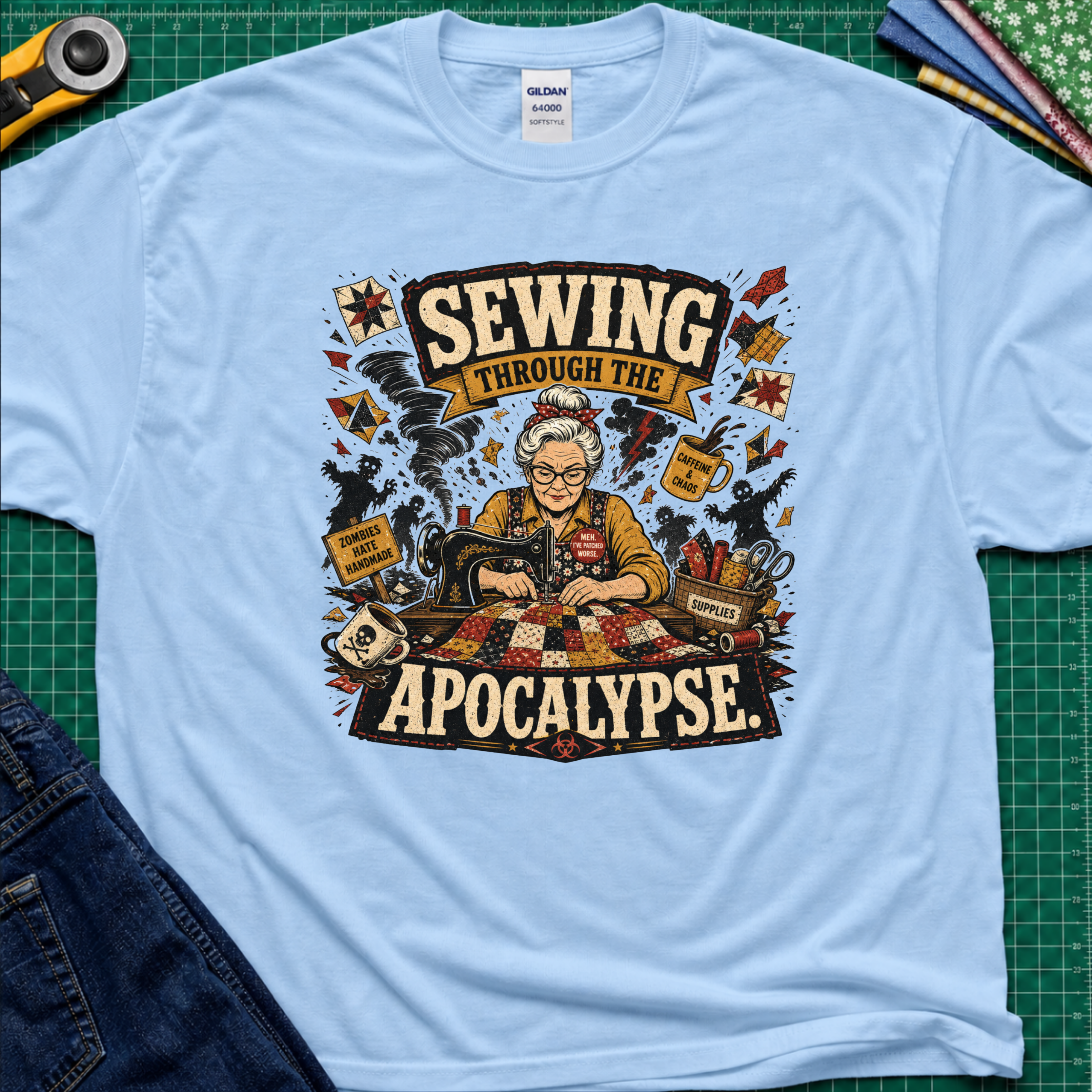 A light blue Gildan t-shirt laid flat on a green cutting mat, featuring a detailed graphic of a woman sewing with the text 'SEWING THROUGH THE APOCALYPSE' and various retro sewing and apocalypse-themed illustrations.